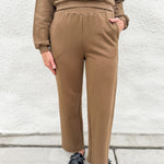 A person wearing a matching brown lounge set consisting of a long-sleeve sweatshirt and wide-leg pants, paired with black loafer clogs, standing against a white textured wall.