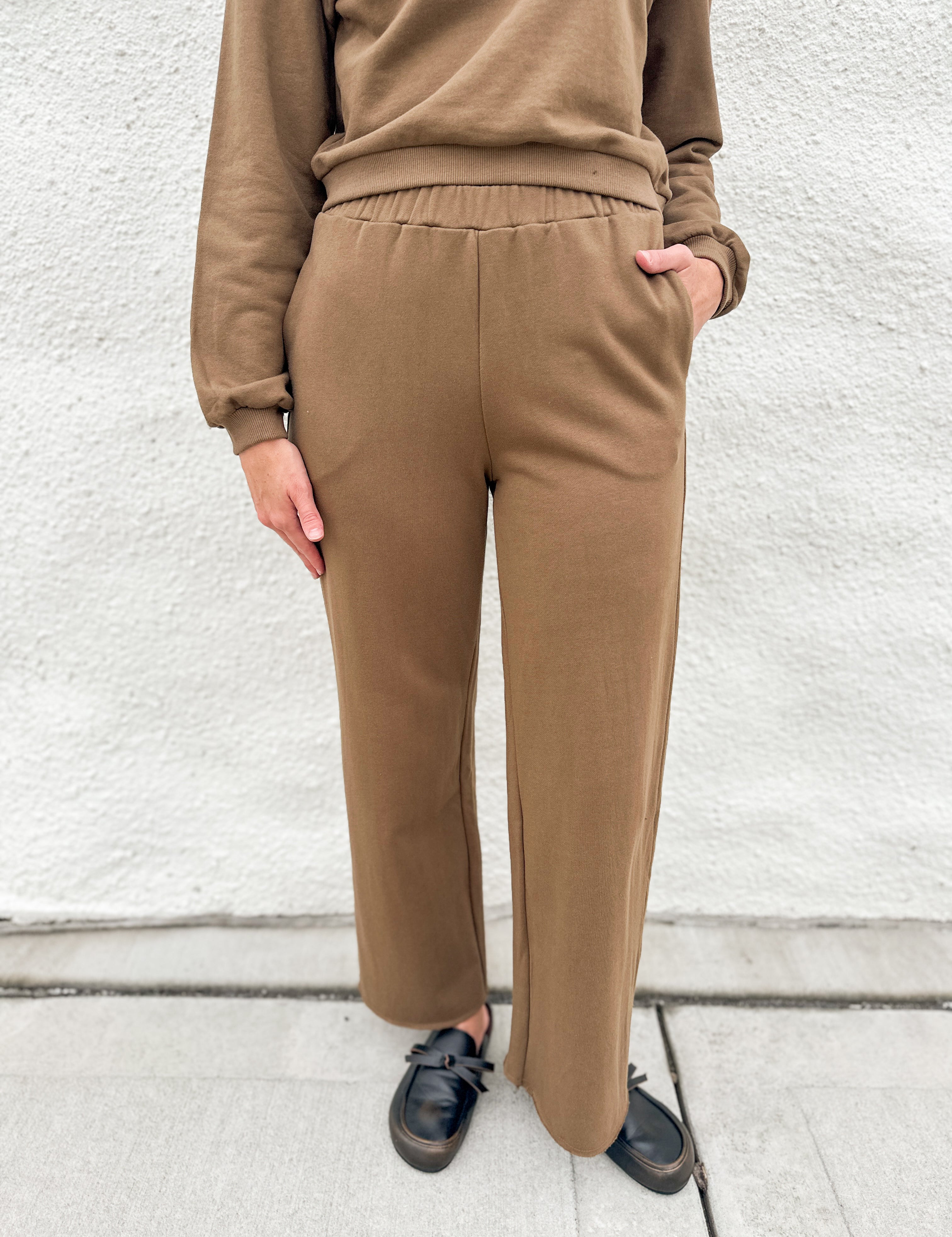 A person wearing a matching brown lounge set consisting of a long-sleeve sweatshirt and wide-leg pants, paired with black loafer clogs, standing against a white textured wall.