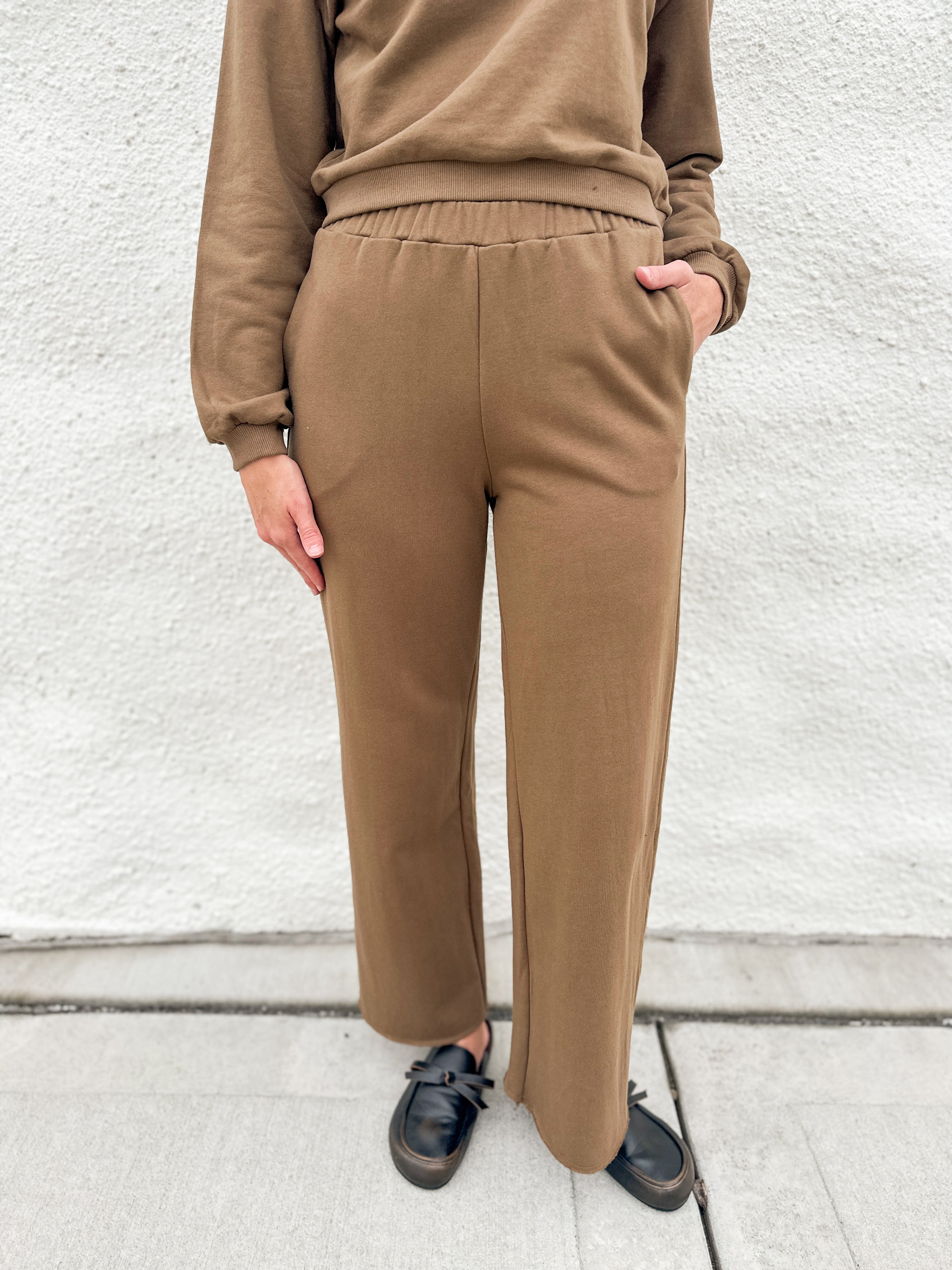A person wearing a matching brown lounge set consisting of a long-sleeve sweatshirt and wide-leg pants, paired with black loafer clogs, standing against a white textured wall.