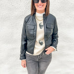 A woman wearing a black faux-leather moto jacket over a cream knit sweater with skull embroidery, paired with gray jeans and black sunglasses, standing against a white textured wall.