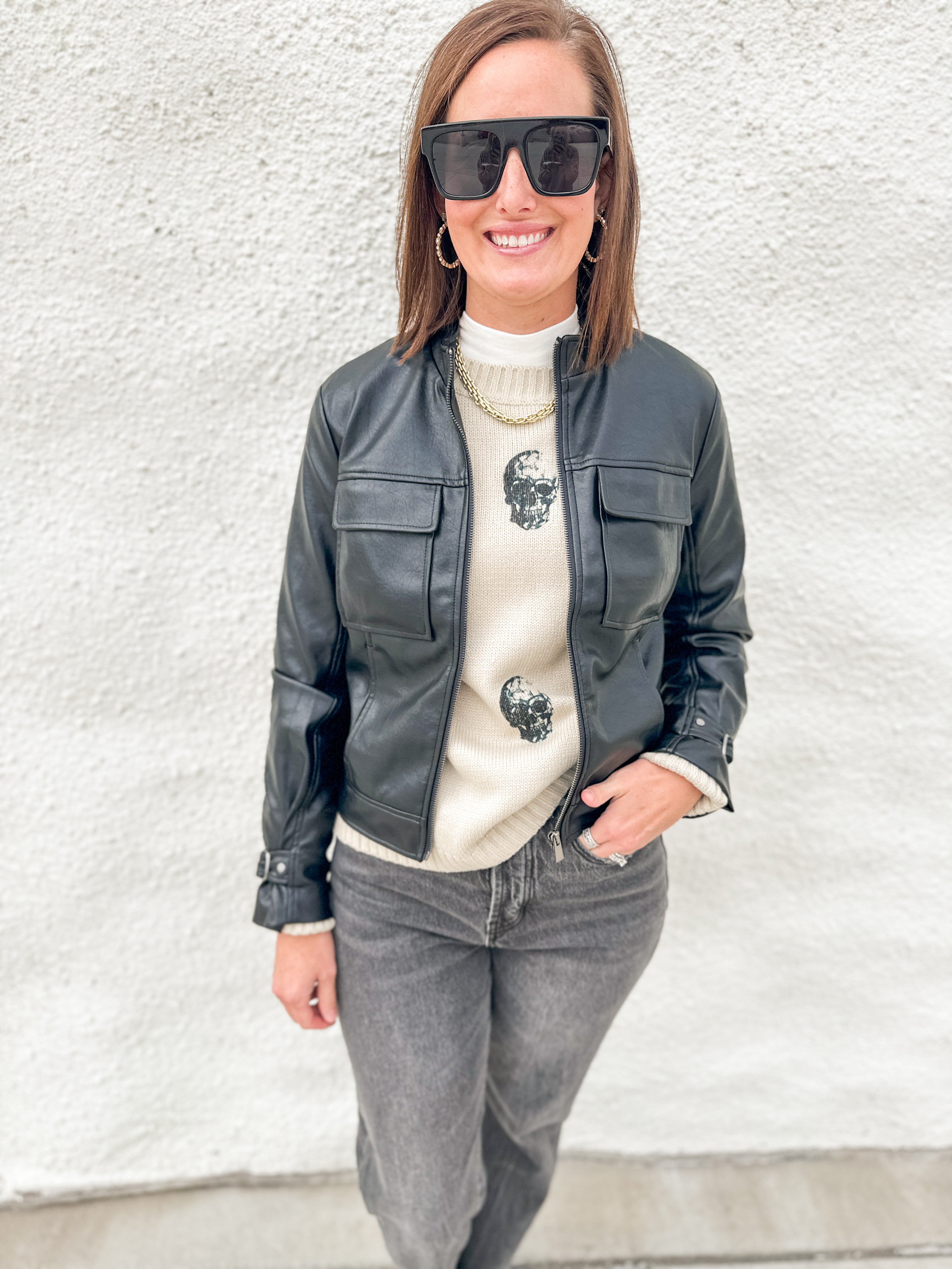 A woman wearing a black faux-leather moto jacket over a cream knit sweater with skull embroidery, paired with gray jeans and black sunglasses, standing against a white textured wall.