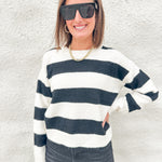 A woman wearing a black and white horizontally striped knit sweater, black sunglasses, and a gold chain necklace, standing against a textured white stucco wall.