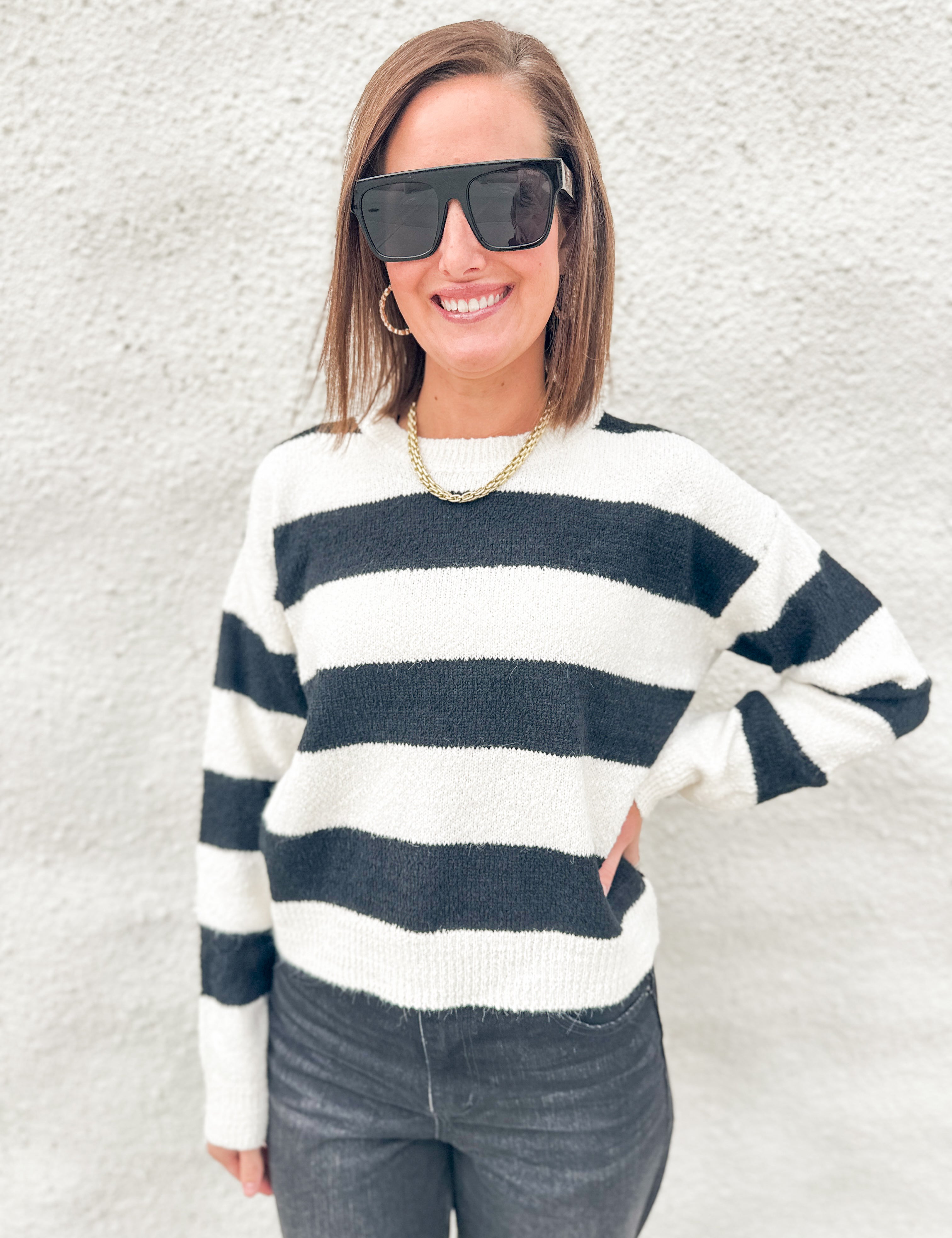 A woman wearing a black and white horizontally striped knit sweater, black sunglasses, and a gold chain necklace, standing against a textured white stucco wall.