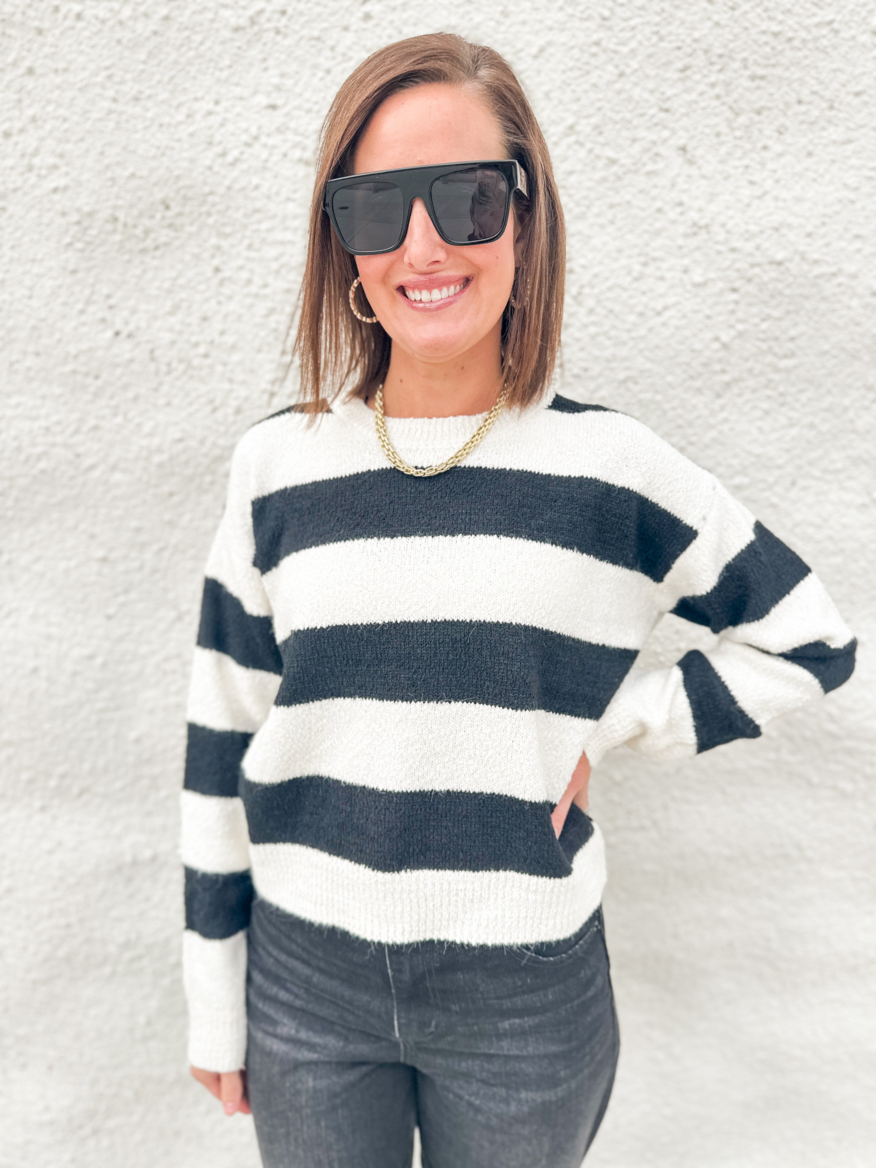 A woman wearing a black and white horizontally striped knit sweater, black sunglasses, and a gold chain necklace, standing against a textured white stucco wall.