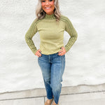 A woman wearing a green thermal top with cable knit sweater sleeves wearing a blue straight leg jean with seaming details down the side and snake print mule clogs standing against a white wall.