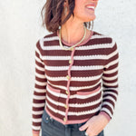 A woman wearing a brown and cream striped knit cardigan with pink trim and gold buttons, paired with dark gray jeans, standing against a textured white wall.