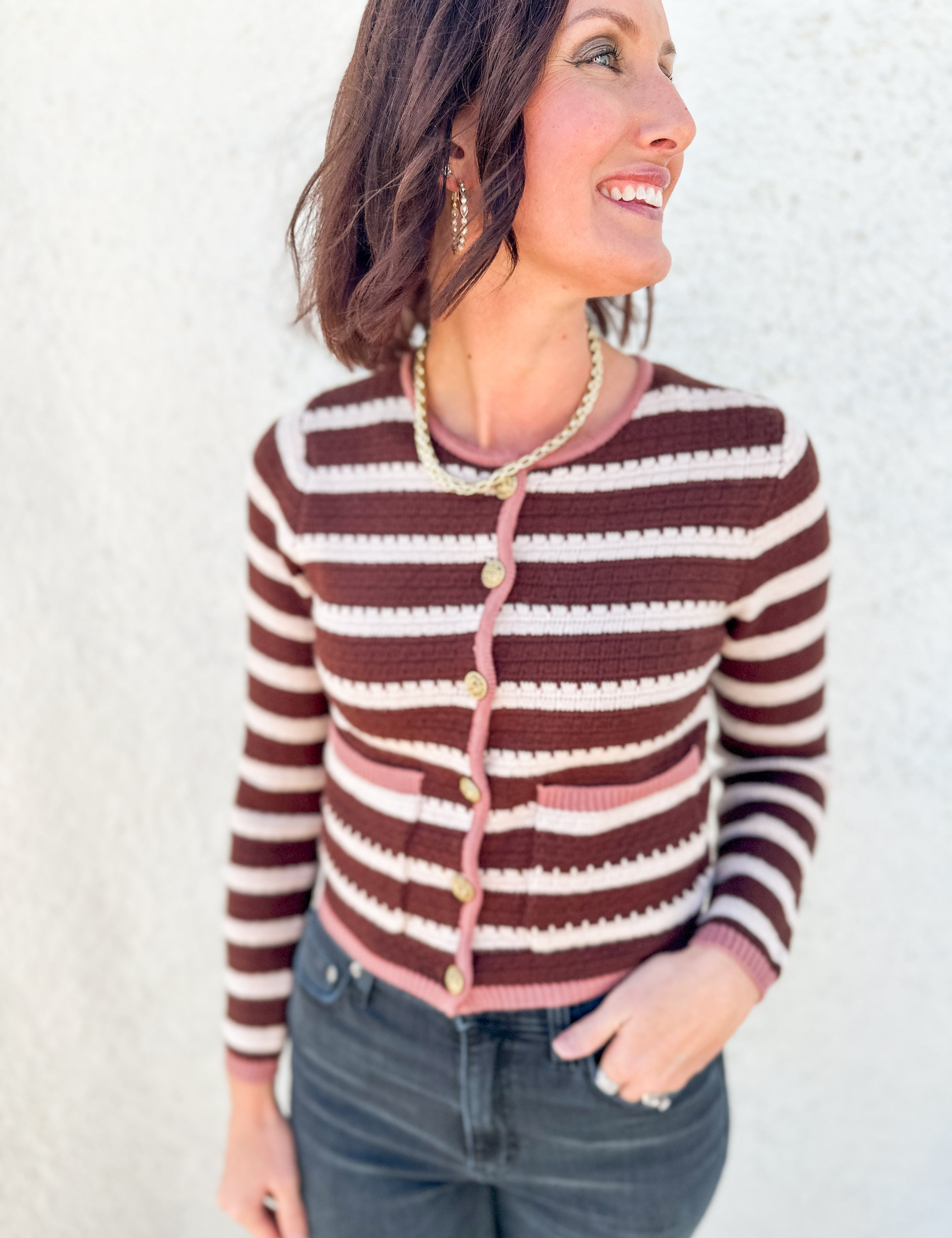 A woman wearing a brown and cream striped knit cardigan with pink trim and gold buttons, paired with dark gray jeans, standing against a textured white wall.