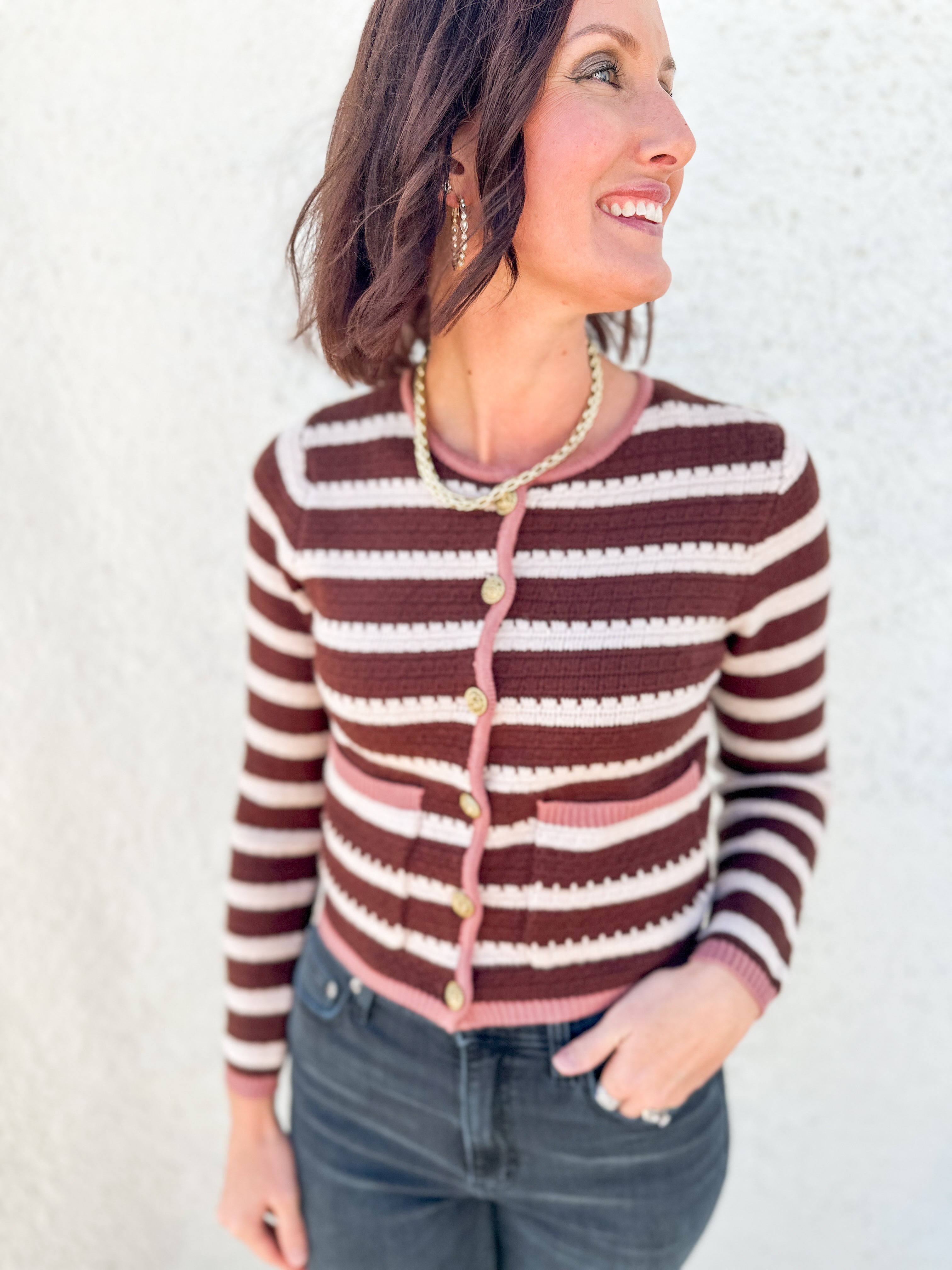 A woman wearing a brown and cream striped knit cardigan with pink trim and gold buttons, paired with dark gray jeans, standing against a textured white wall.