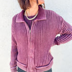 A woman wearing a ribbed knit zip-front cardigan in dusty mauve pink, paired with a dark purple top and dark gray jeans, standing against a textured white wall.