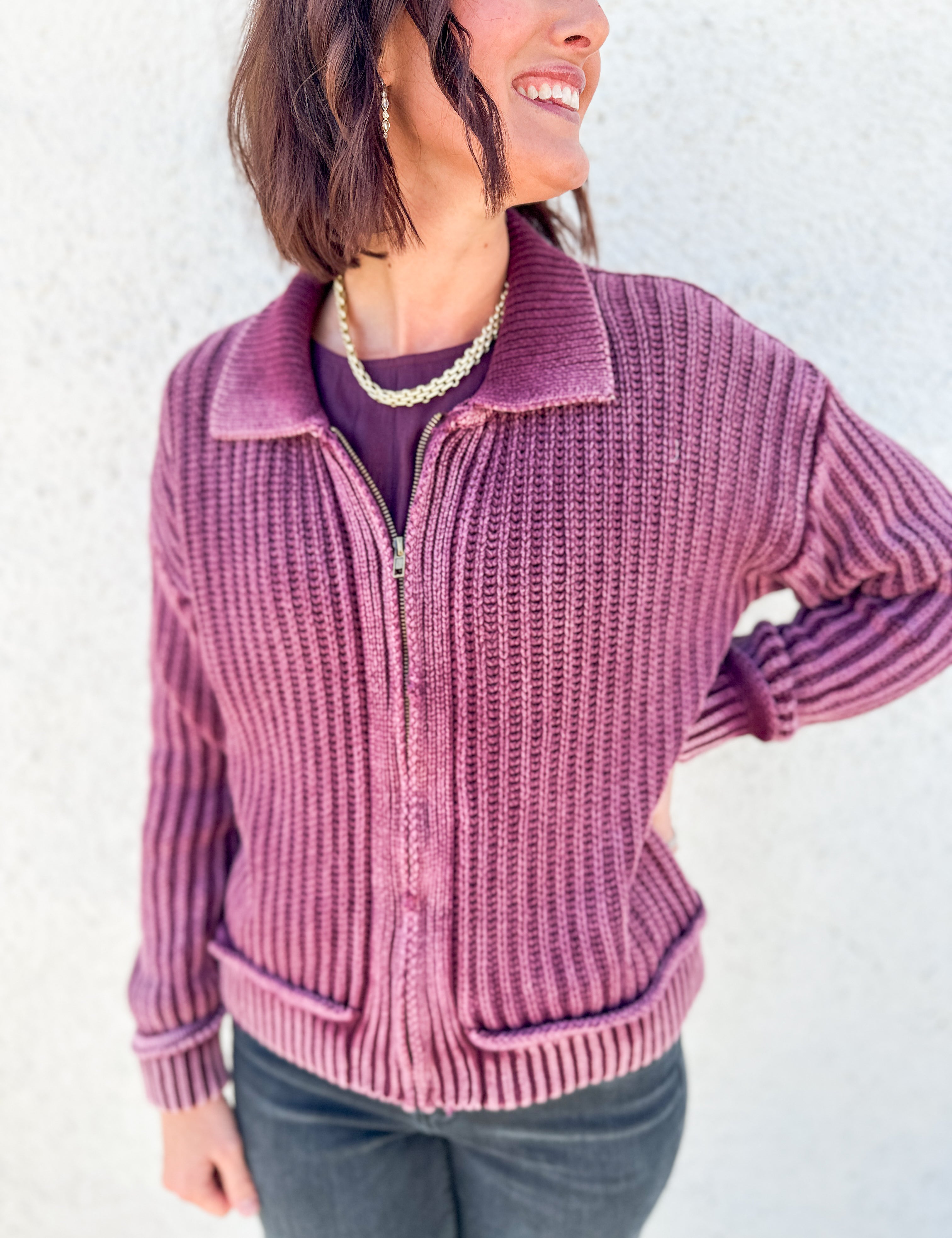 A woman wearing a ribbed knit zip-front cardigan in dusty mauve pink, paired with a dark purple top and dark gray jeans, standing against a textured white wall.
