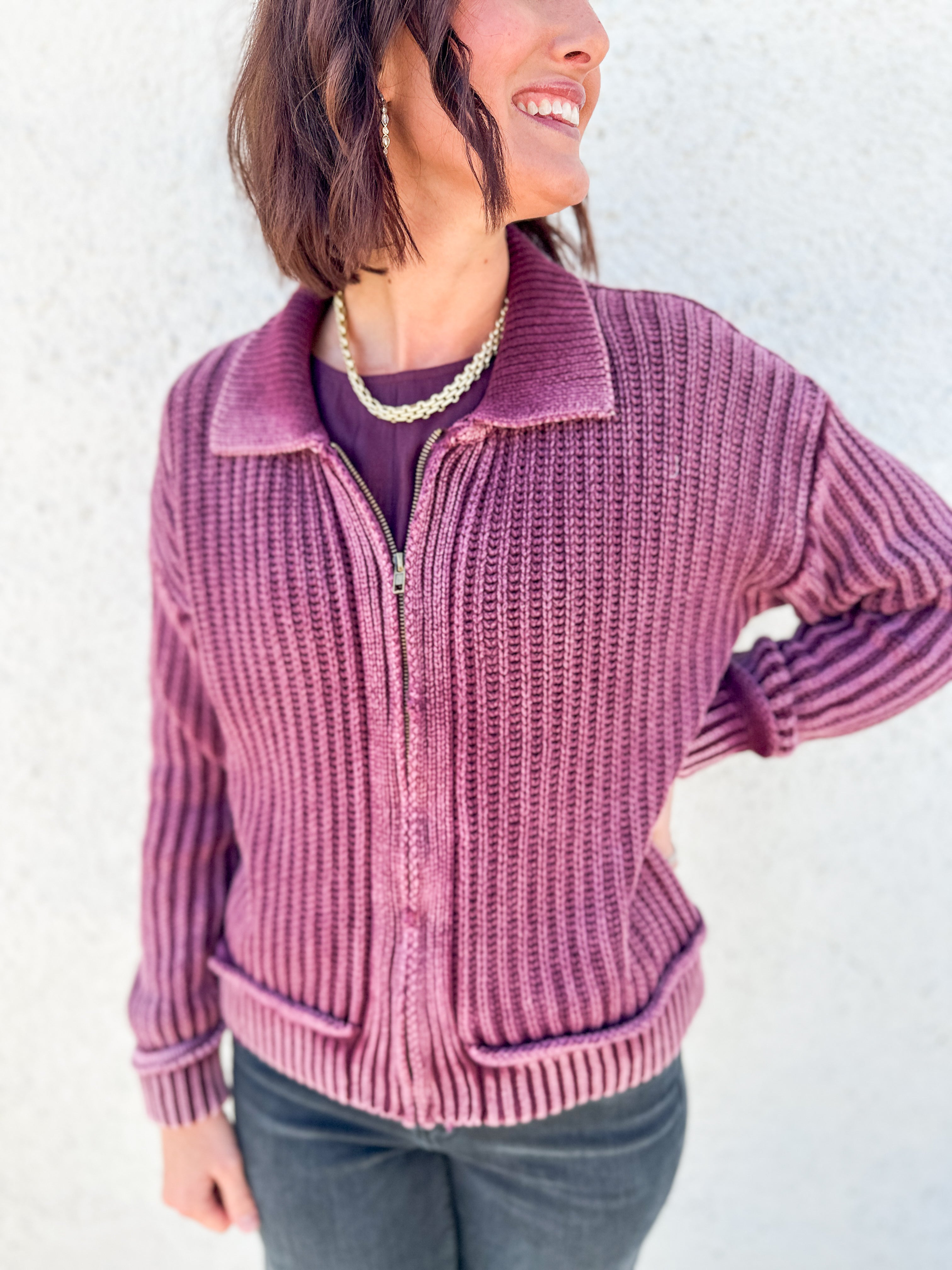 A woman wearing a ribbed knit zip-front cardigan in dusty mauve pink, paired with a dark purple top and dark gray jeans, standing against a textured white wall.