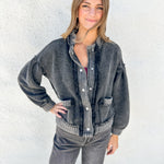 A woman wearing a faded black washed denim button-up shirt jacket with ribbed trim and distressed details, paired with gray jeans, standing against a white stucco wall.