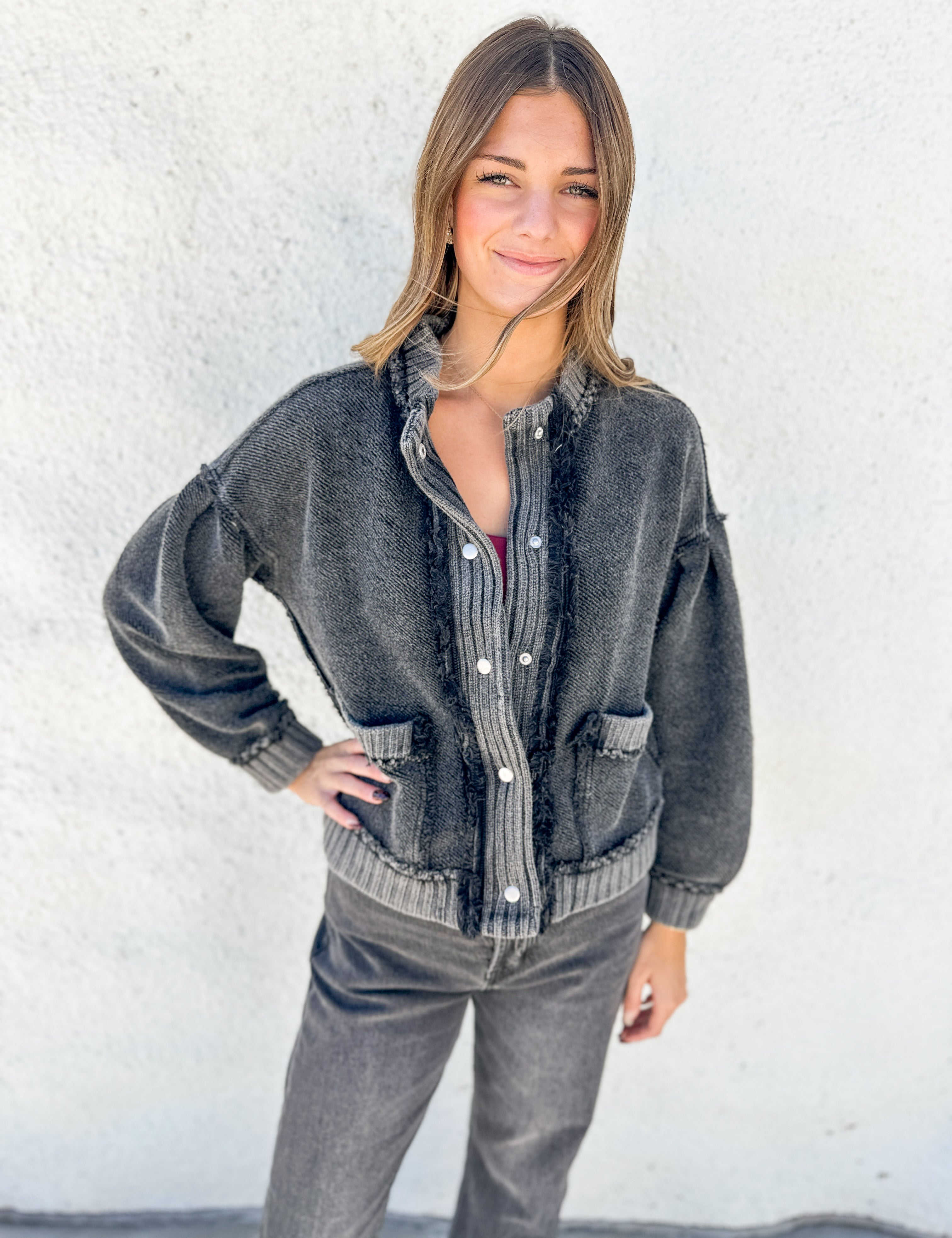 A woman wearing a faded black washed denim button-up shirt jacket with ribbed trim and distressed details, paired with gray jeans, standing against a white stucco wall.
