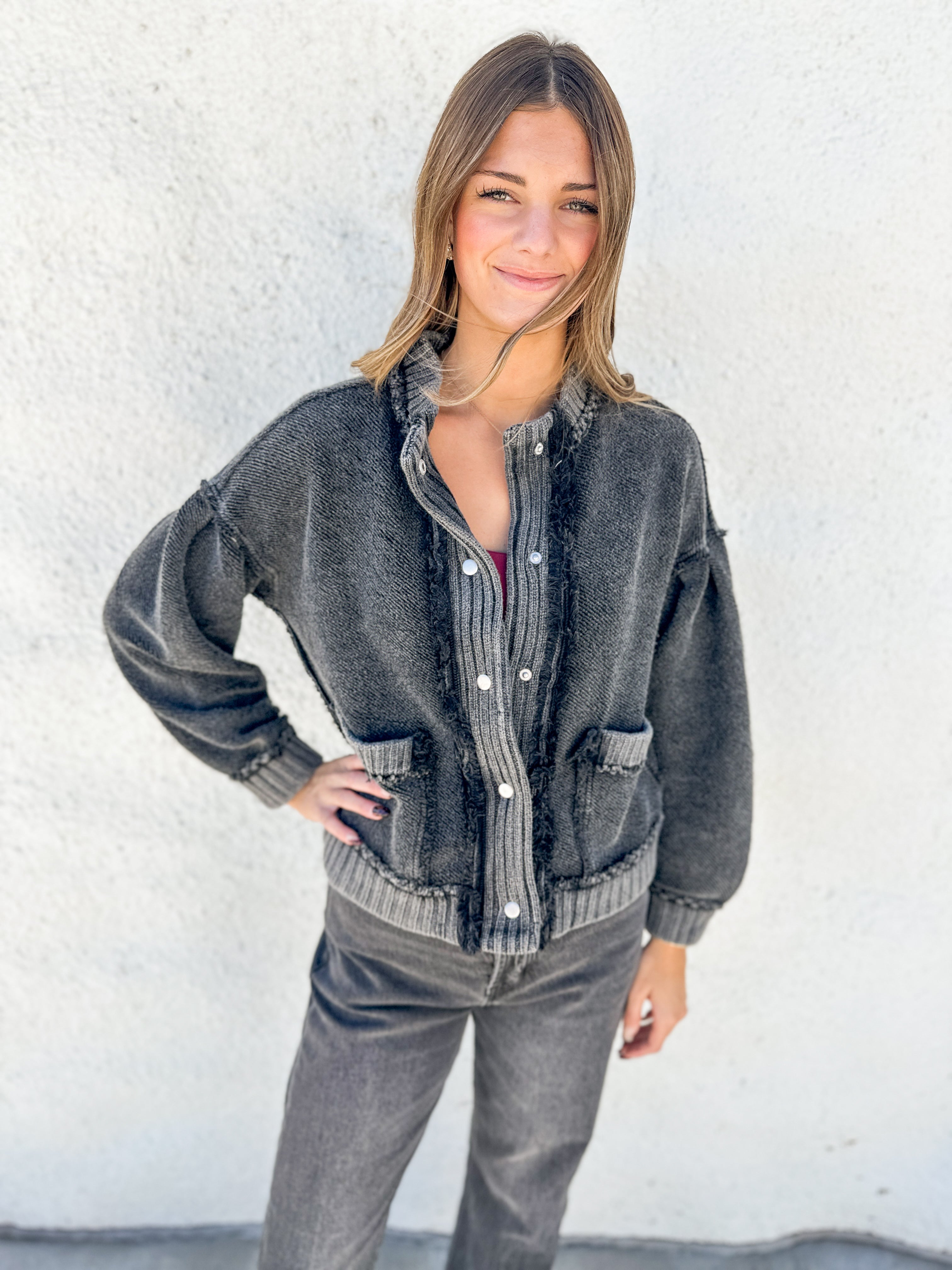 A woman wearing a faded black washed denim button-up shirt jacket with ribbed trim and distressed details, paired with gray jeans, standing against a white stucco wall.