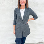 A woman wearing a gray and black plaid sweater blazer over a white top and dark pants, standing against a white textured wall.