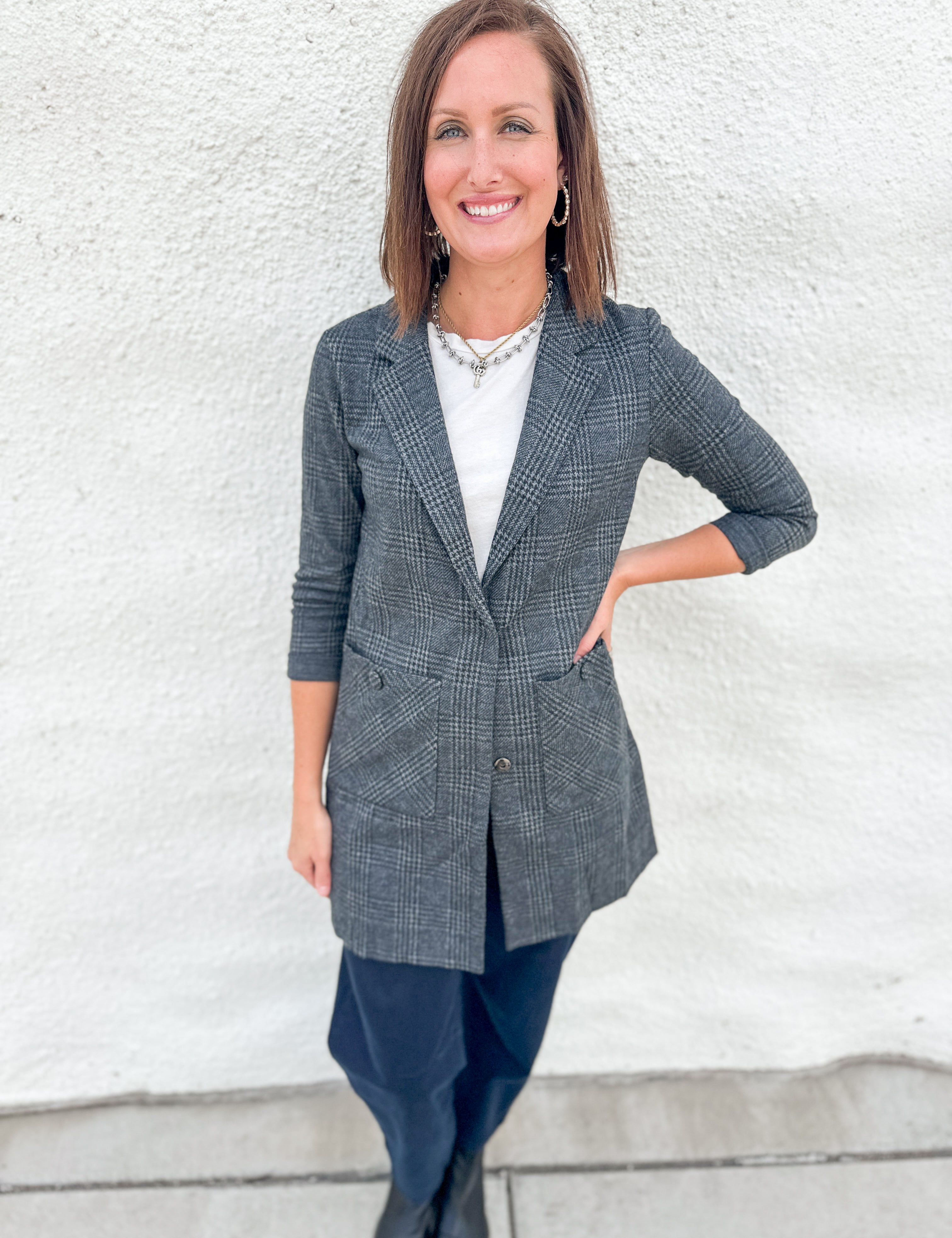 A woman wearing a gray and black plaid sweater blazer over a white top and dark pants, standing against a white textured wall.