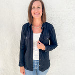 A woman smiling and posing in front of a textured white wall, wearing a black button-up overshirt over a white long sleeve top and light blue jeans.