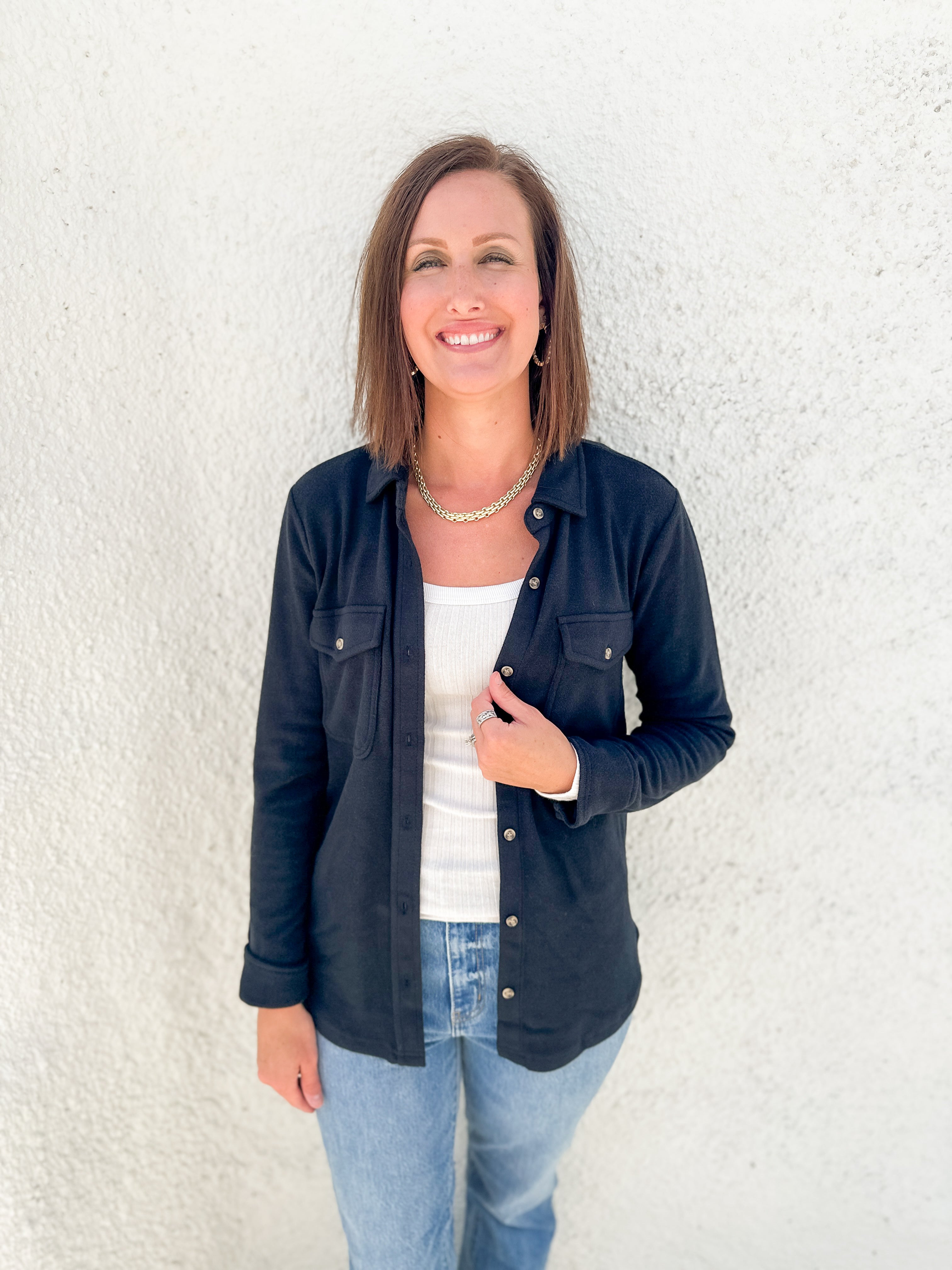 A woman smiling and posing in front of a textured white wall, wearing a black button-up overshirt over a white long sleeve top and light blue jeans.