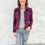 A woman wearing a stretchy burgundy utility jacket with military band jacket styling and peplum back over a gray patterned top and light-wash jeans, standing against a white textured wall.