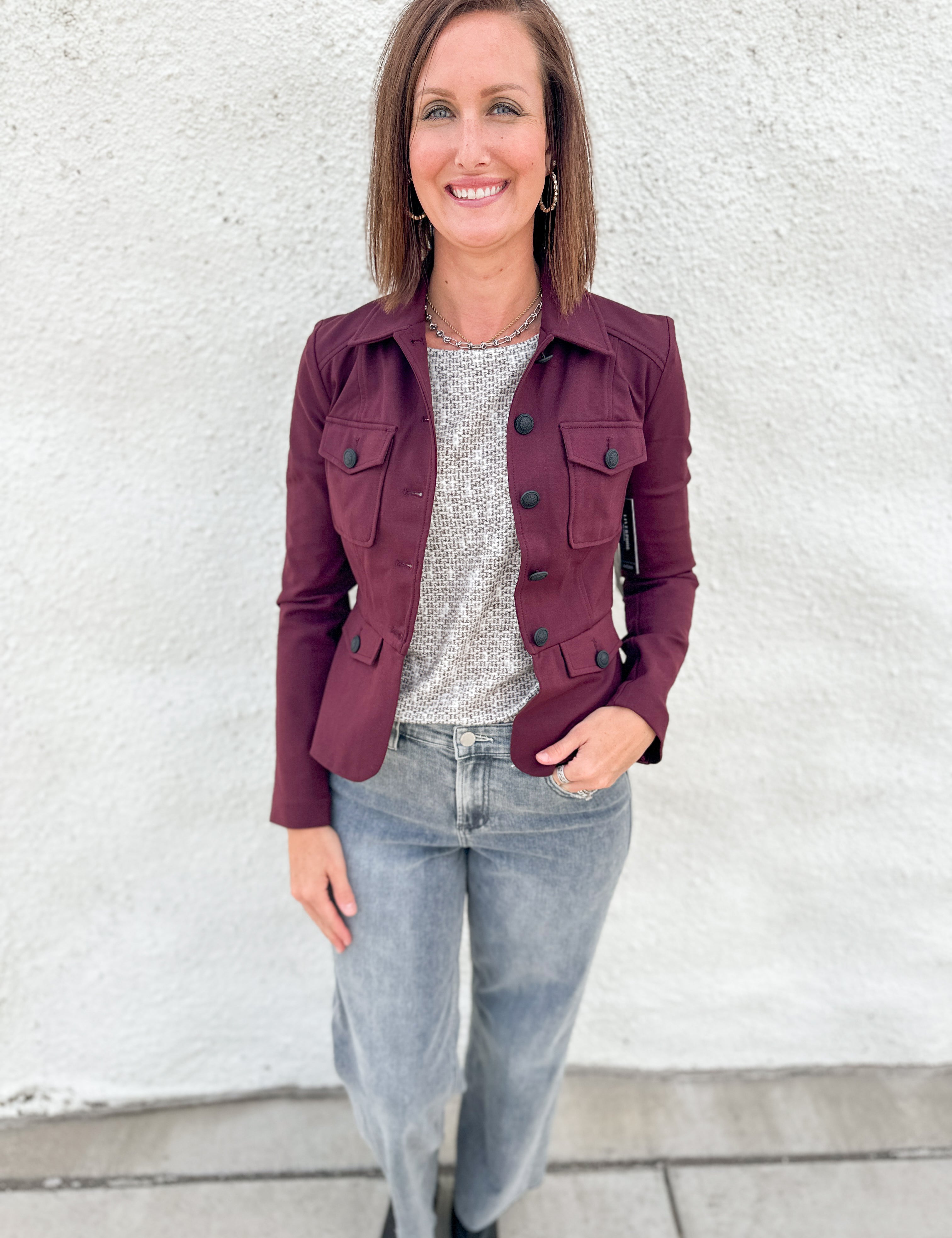 A woman wearing a stretchy burgundy utility jacket with military band jacket styling and peplum back over a gray patterned top and light-wash jeans, standing against a white textured wall.