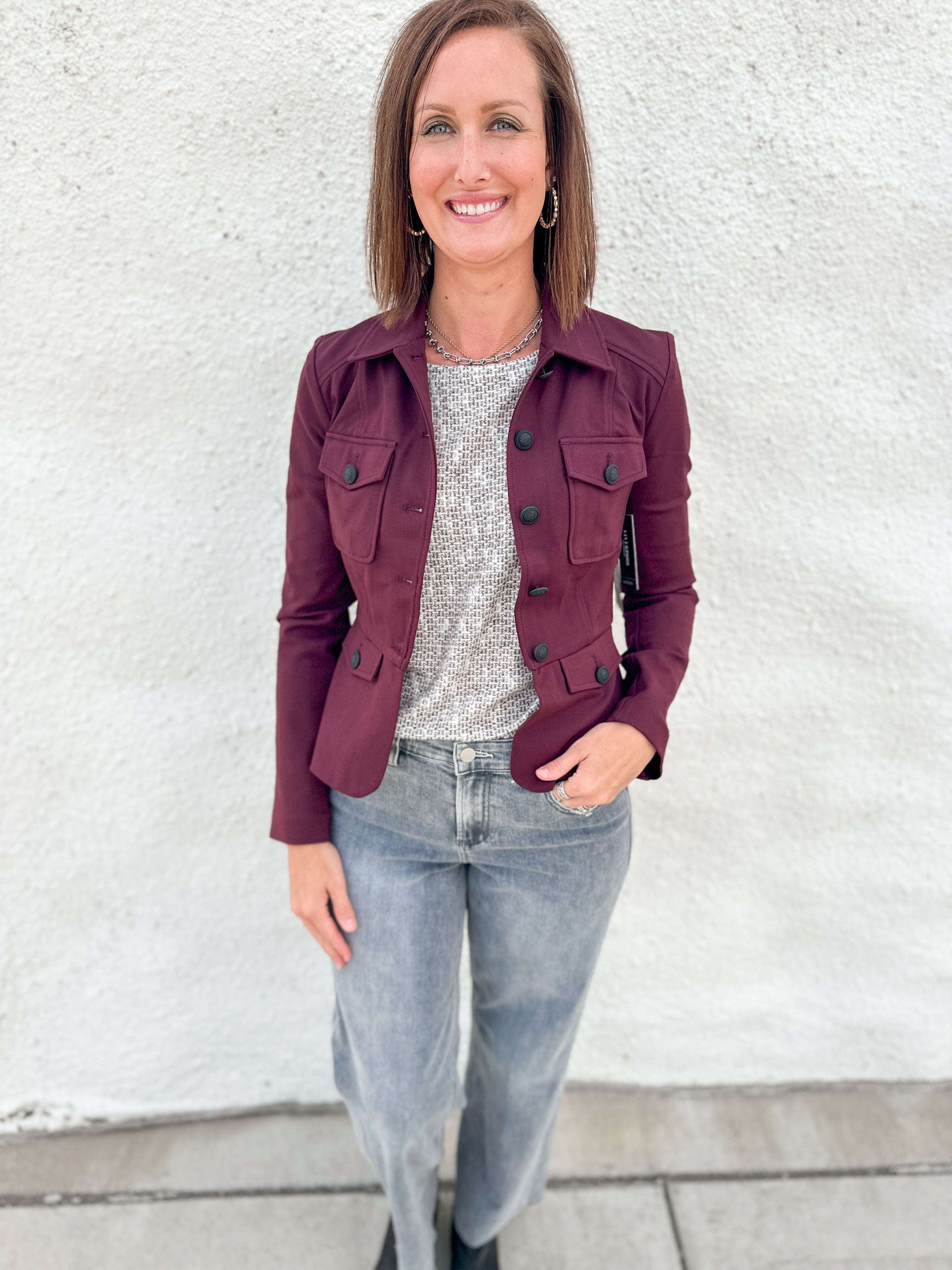 A woman wearing a stretchy burgundy utility jacket with military band jacket styling and peplum back over a gray patterned top and light-wash jeans, standing against a white textured wall.