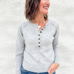 A woman wearing a light gray knit henley sweater with brown buttons, paired with blue jeans, standing against a textured white wall.