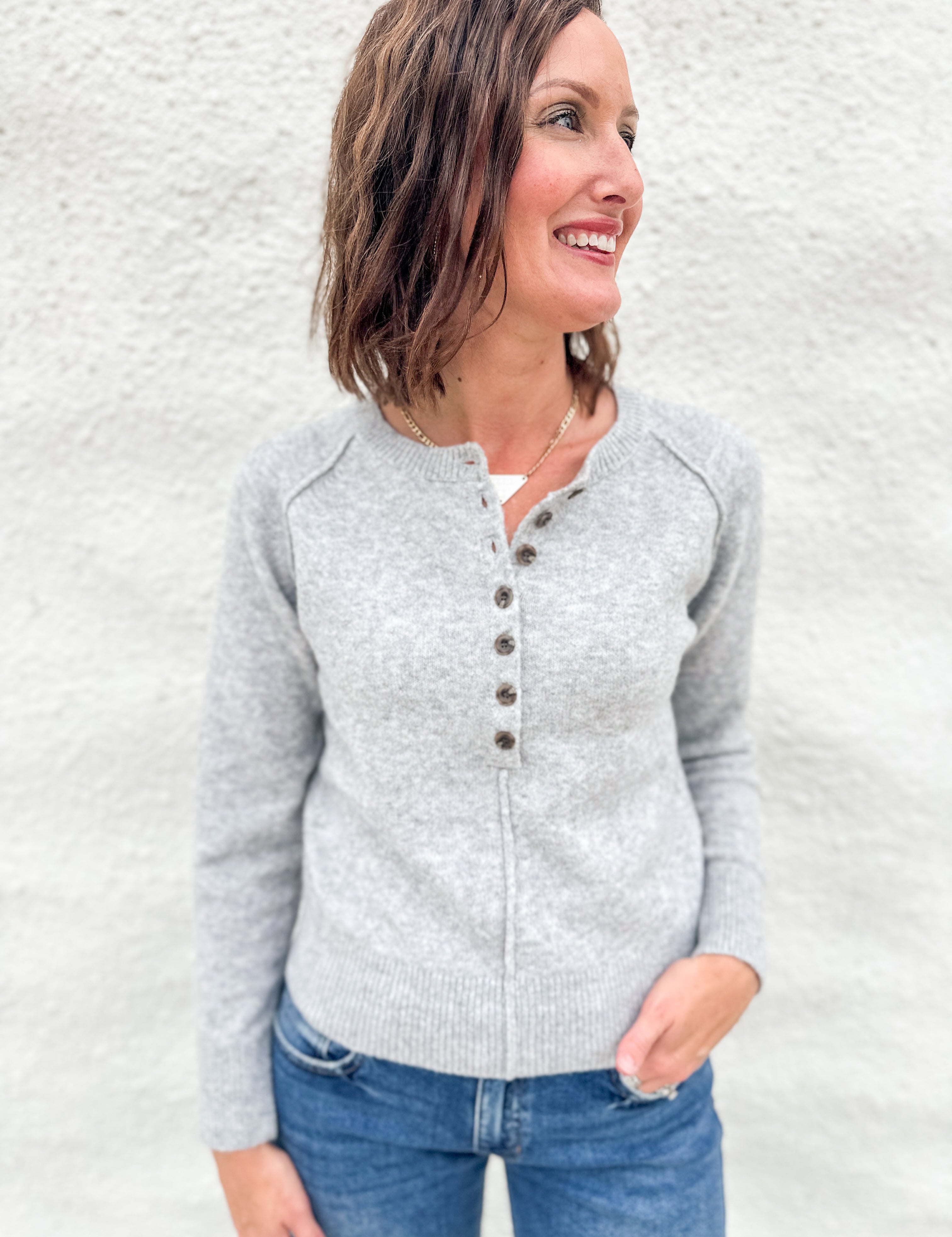 A woman wearing a light gray knit henley sweater with brown buttons, paired with blue jeans, standing against a textured white wall.