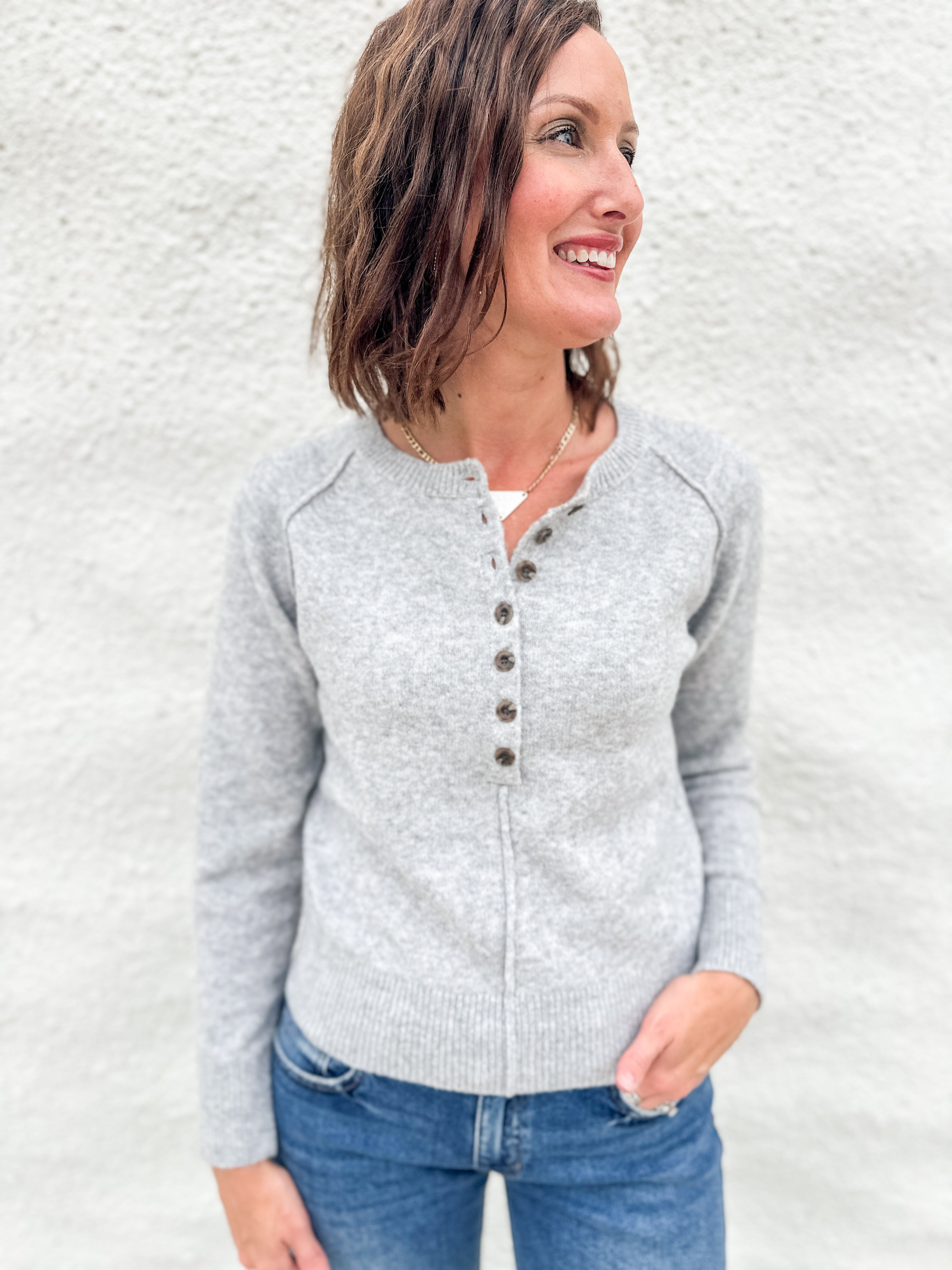 A woman wearing a light gray knit henley sweater with brown buttons, paired with blue jeans, standing against a textured white wall.