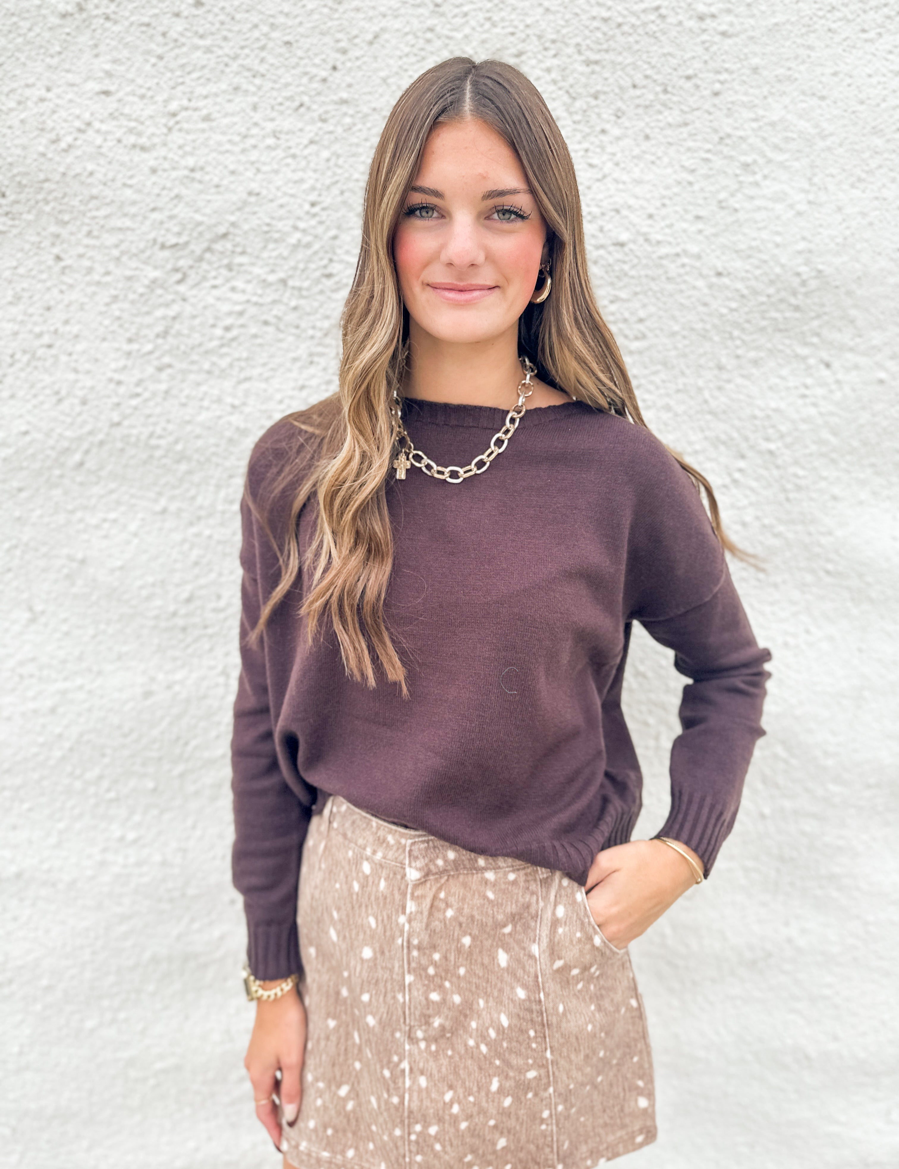 A woman modeling a dark chocolate colored knit sweater and a beige fawn print denim mini skirt against a textured white wall.