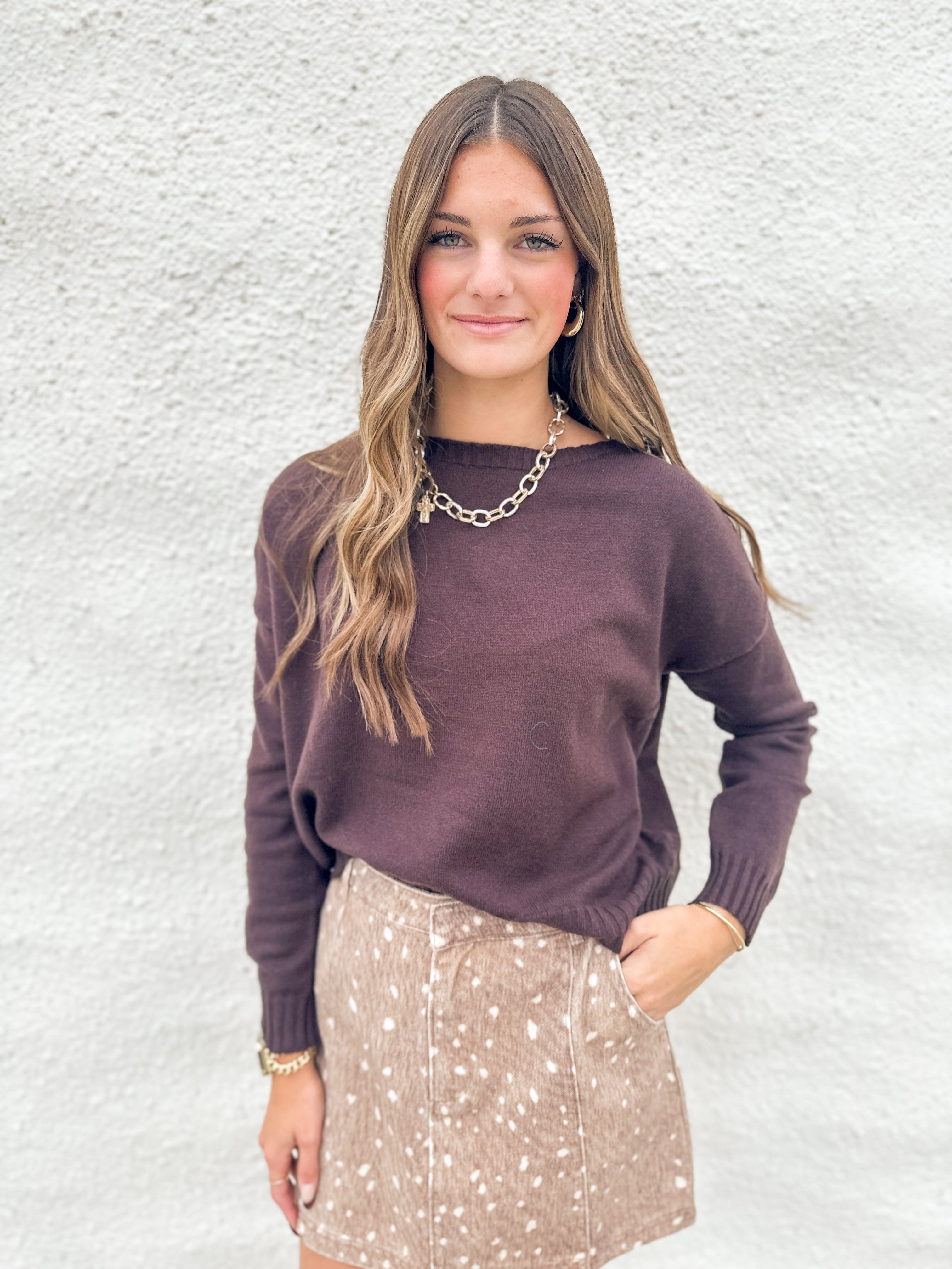 A woman modeling a dark chocolate colored knit sweater and a beige fawn print denim mini skirt against a textured white wall.