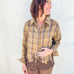 A woman wearing a brown and tan plaid button-up shirt with brown lace trim at the hem, paired with brown pants, standing against a textured white stucco wall.