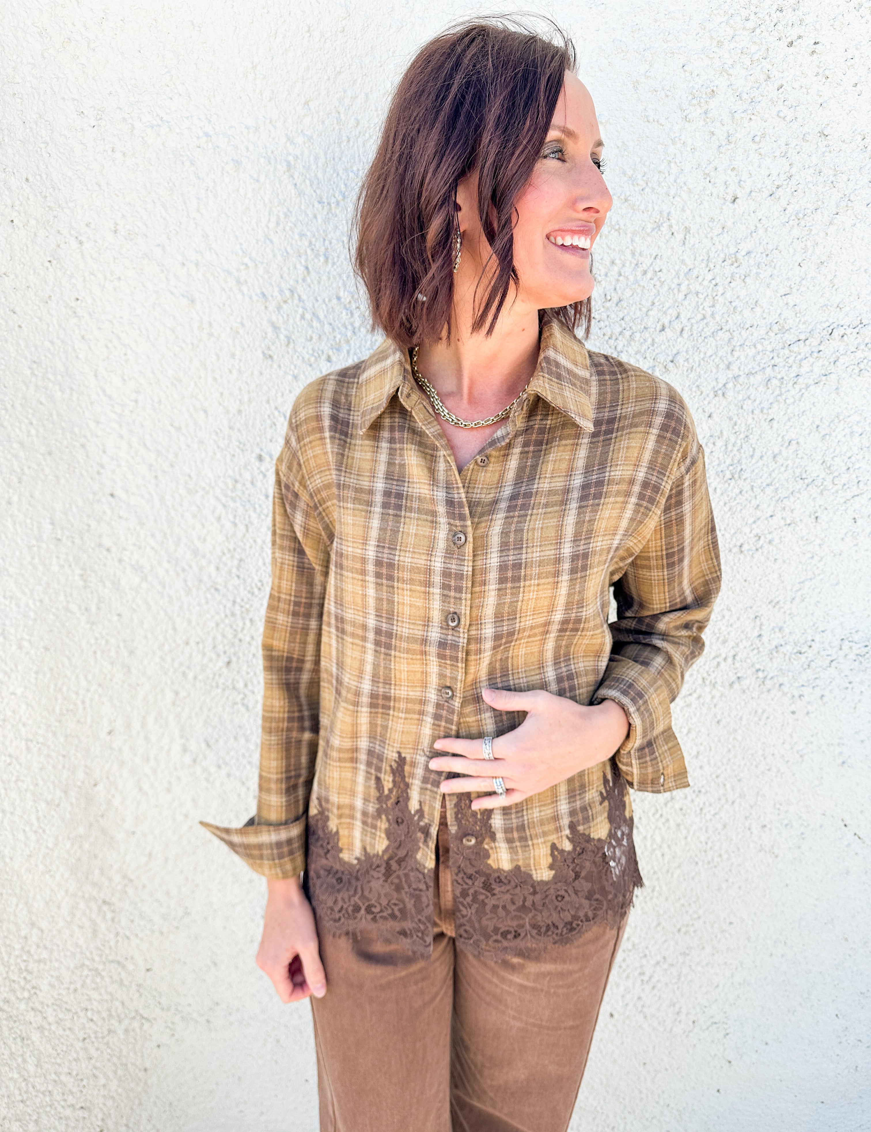 A woman wearing a brown and tan plaid button-up shirt with brown lace trim at the hem, paired with brown pants, standing against a textured white stucco wall.