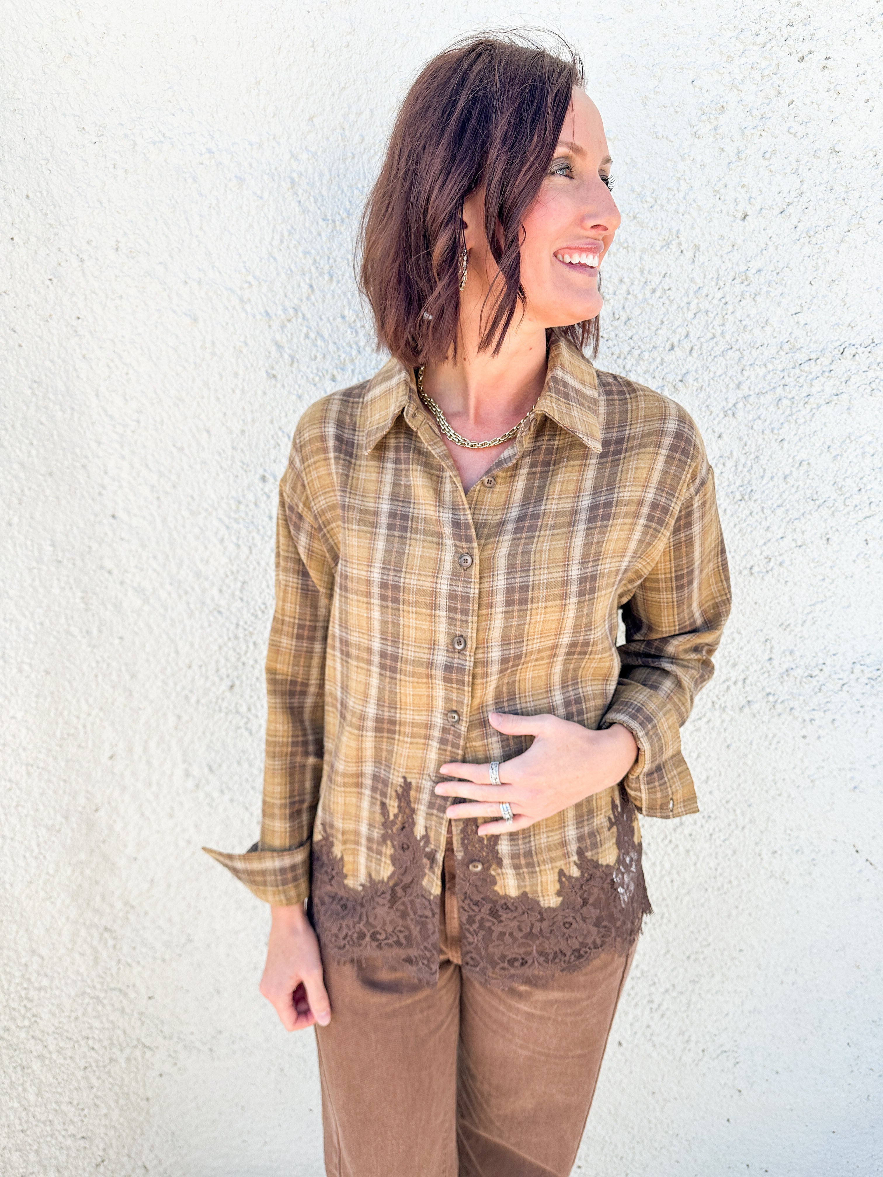 A woman wearing a brown and tan plaid button-up shirt with brown lace trim at the hem, paired with brown pants, standing against a textured white stucco wall.