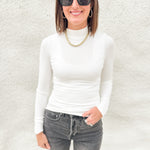 A woman wearing a fitted white lightly ribbed long-sleeve mock neck top, dark gray jeans, black sunglasses, and a gold chain necklace, standing against a textured white wall.