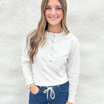 A smiling young woman with long light brown hair stands against a white textured wall, wearing a white long-sleeve henley top with gray buttons and dark blue jeans with a white drawstring.