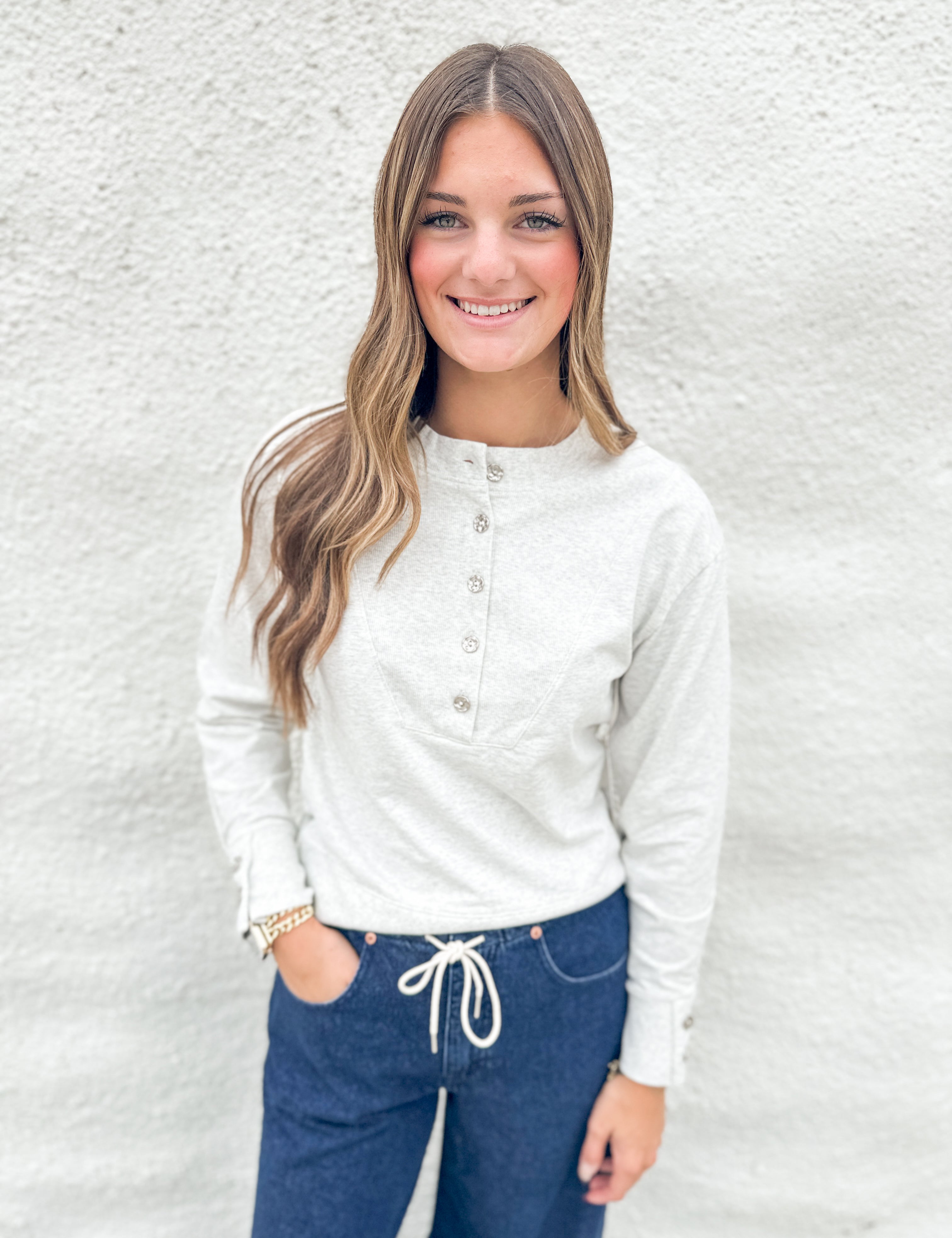 A smiling young woman with long light brown hair stands against a white textured wall, wearing a white long-sleeve henley top with gray buttons and dark blue jeans with a white drawstring.