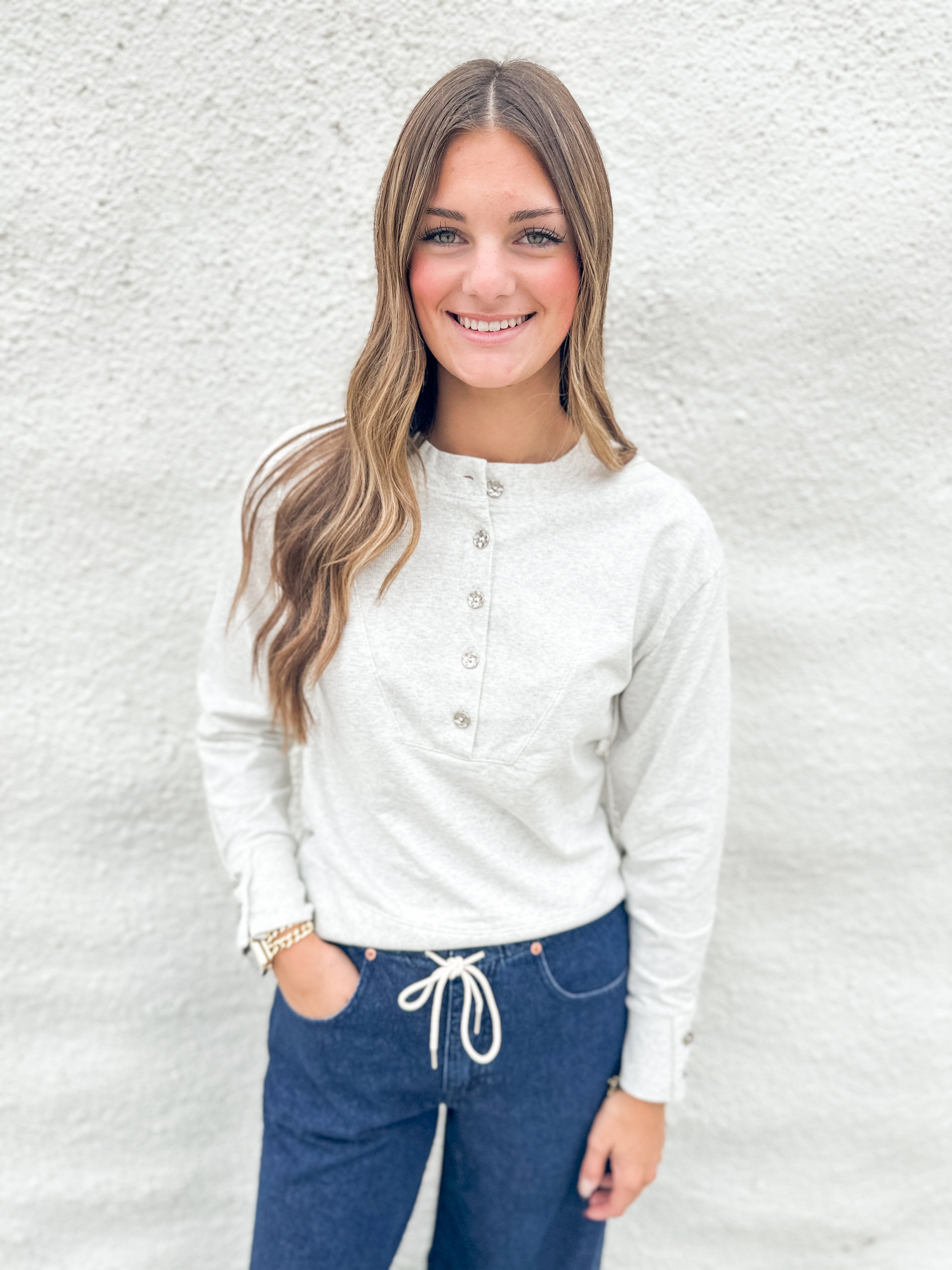 A smiling young woman with long light brown hair stands against a white textured wall, wearing a white long-sleeve henley top with gray buttons and dark blue jeans with a white drawstring.