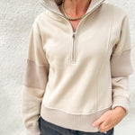 A woman wearing a beige quarter-zip pullover sweater with ribbed trim and contrasting ribbed sleeve panels, paired with dark gray jeans against a textured white wall.