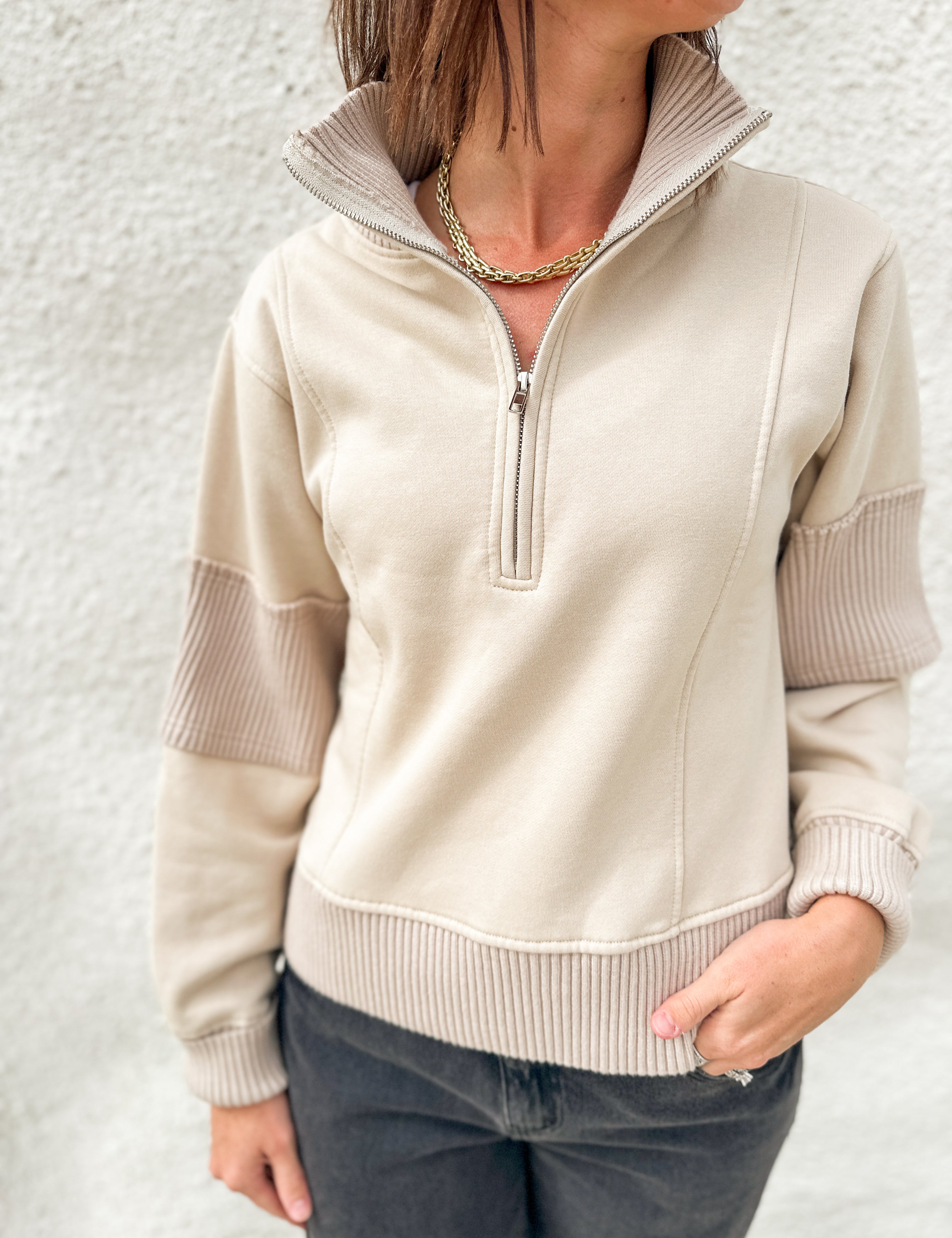 A woman wearing a beige quarter-zip pullover sweater with ribbed trim and contrasting ribbed sleeve panels, paired with dark gray jeans against a textured white wall.
