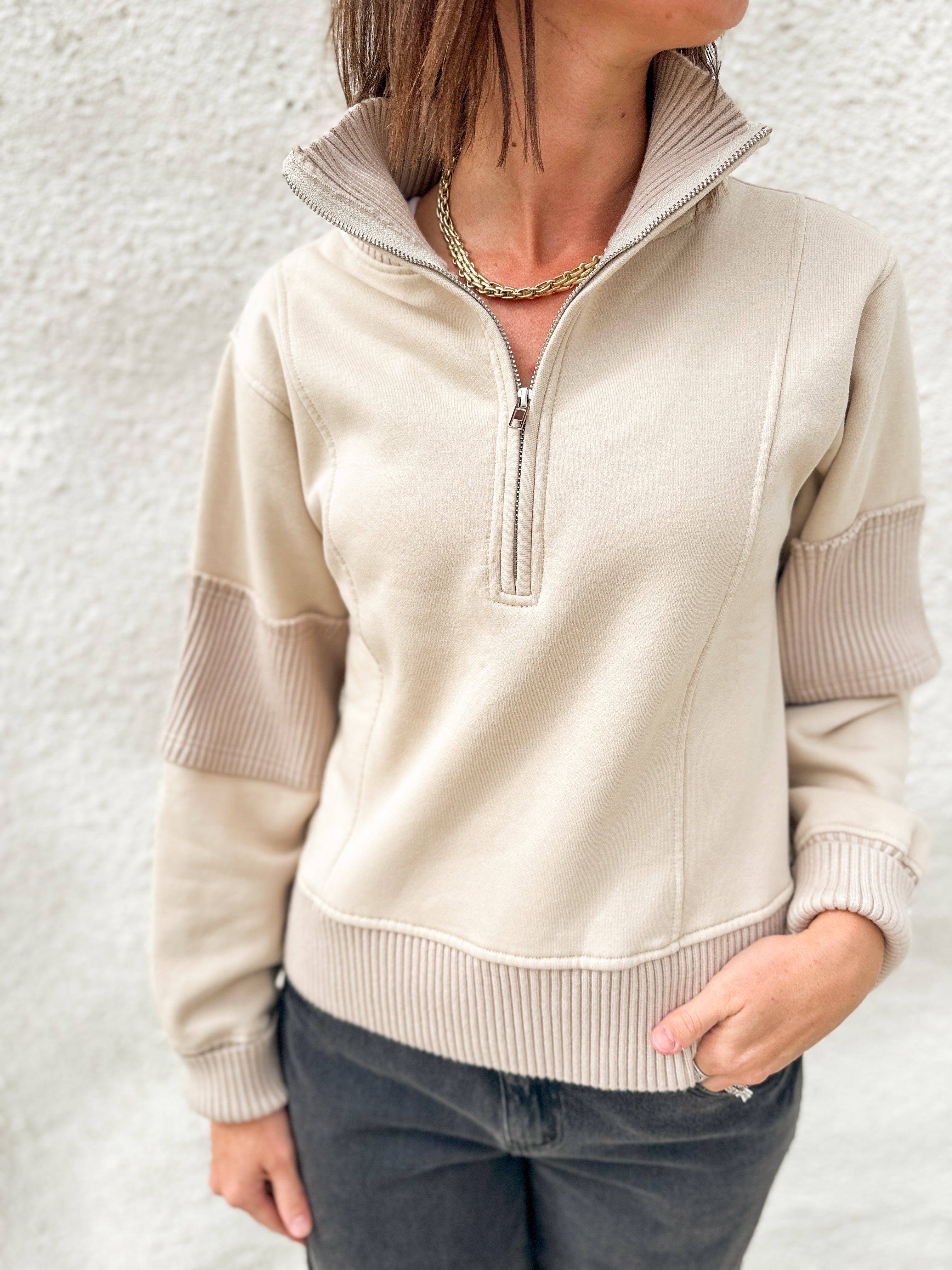 A woman wearing a beige quarter-zip pullover sweater with ribbed trim and contrasting ribbed sleeve panels, paired with dark gray jeans against a textured white wall.