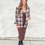 A woman stands outdoors against a white textured wall wearing a brown and beige plaid button-up shirt over a brown midi dress, paired with black pointed ankle boots.