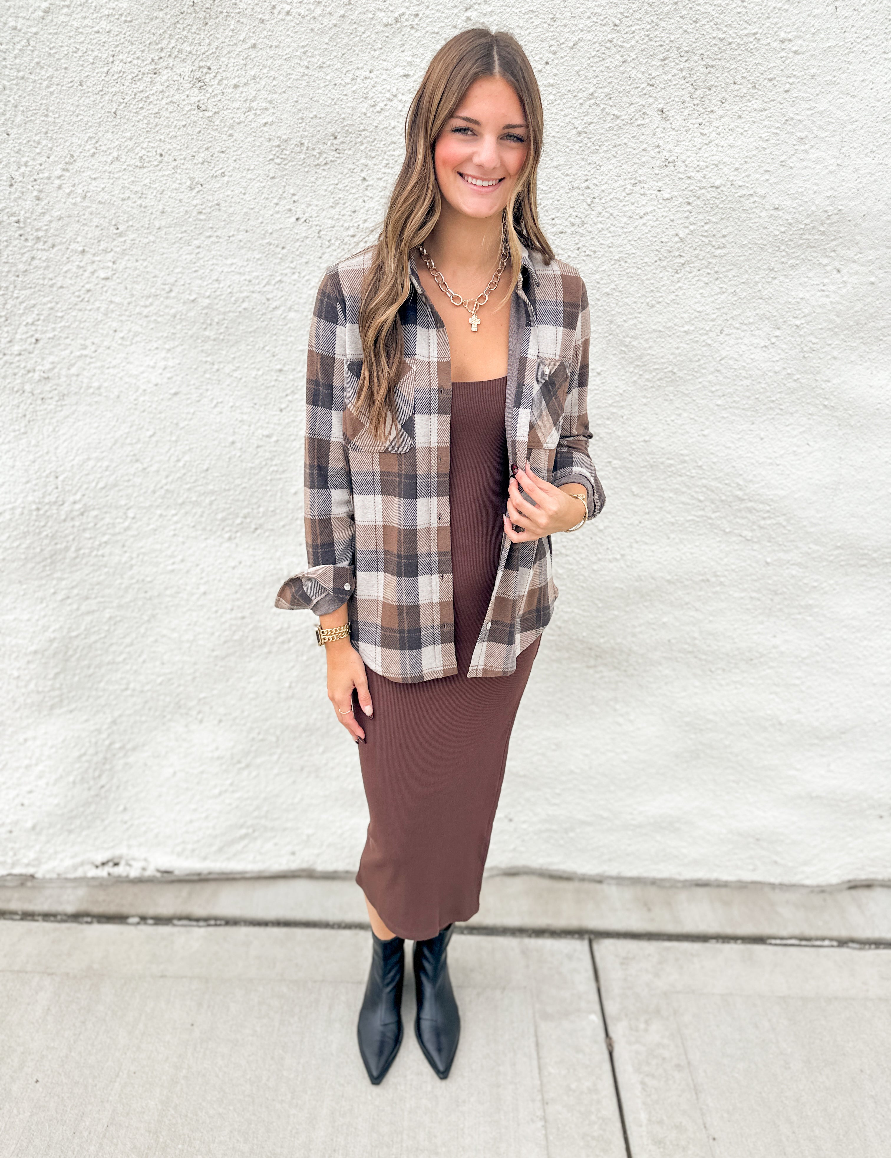 A woman stands outdoors against a white textured wall wearing a brown and beige plaid button-up shirt over a brown midi dress, paired with black pointed ankle boots.
