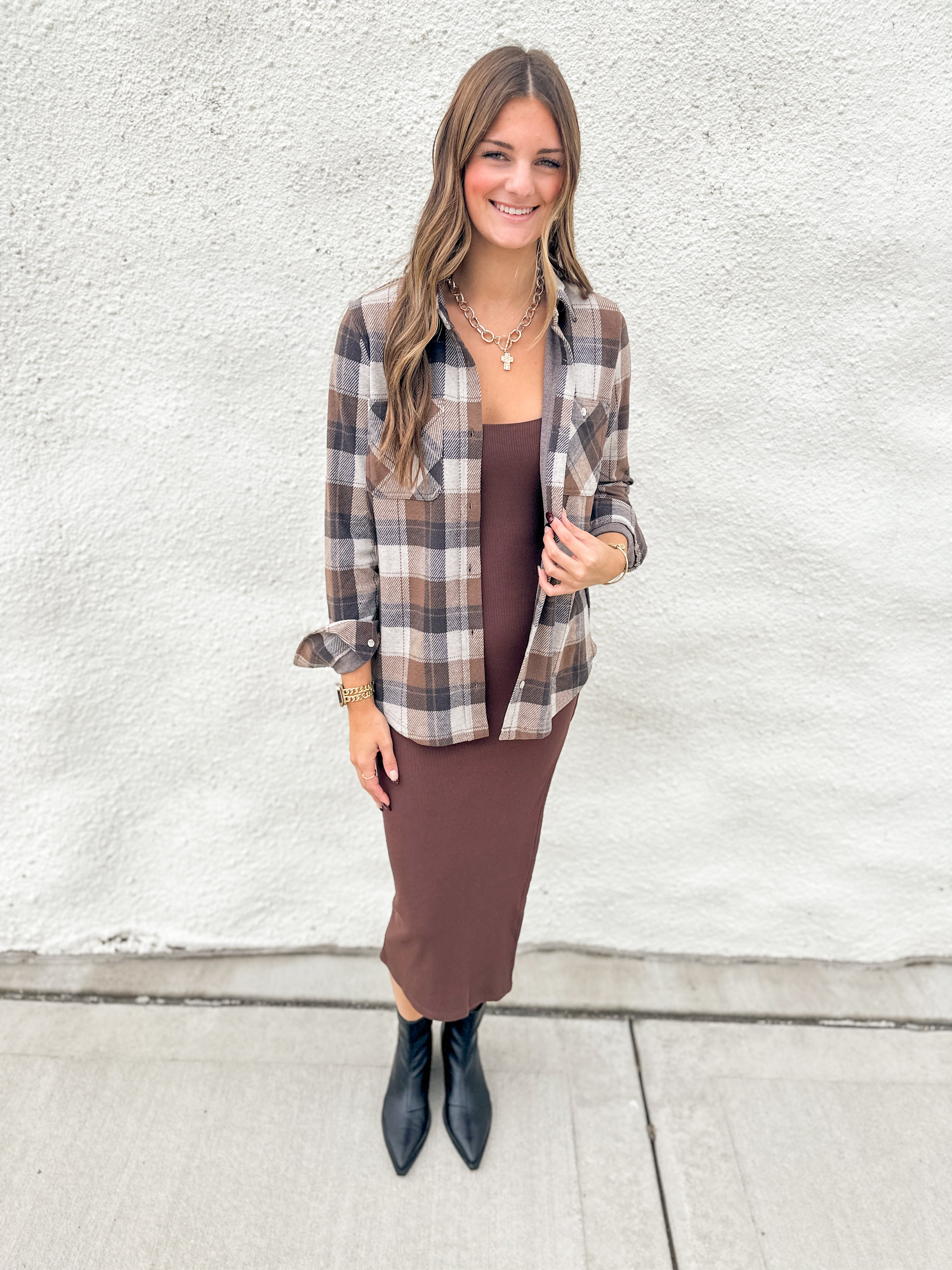 A woman stands outdoors against a white textured wall wearing a brown and beige plaid button-up shirt over a brown midi dress, paired with black pointed ankle boots.
