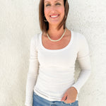 A smiling woman with brown hair stands against a textured white wall, wearing a white long-sleeve ribbed scoop-neck top and blue jeans.