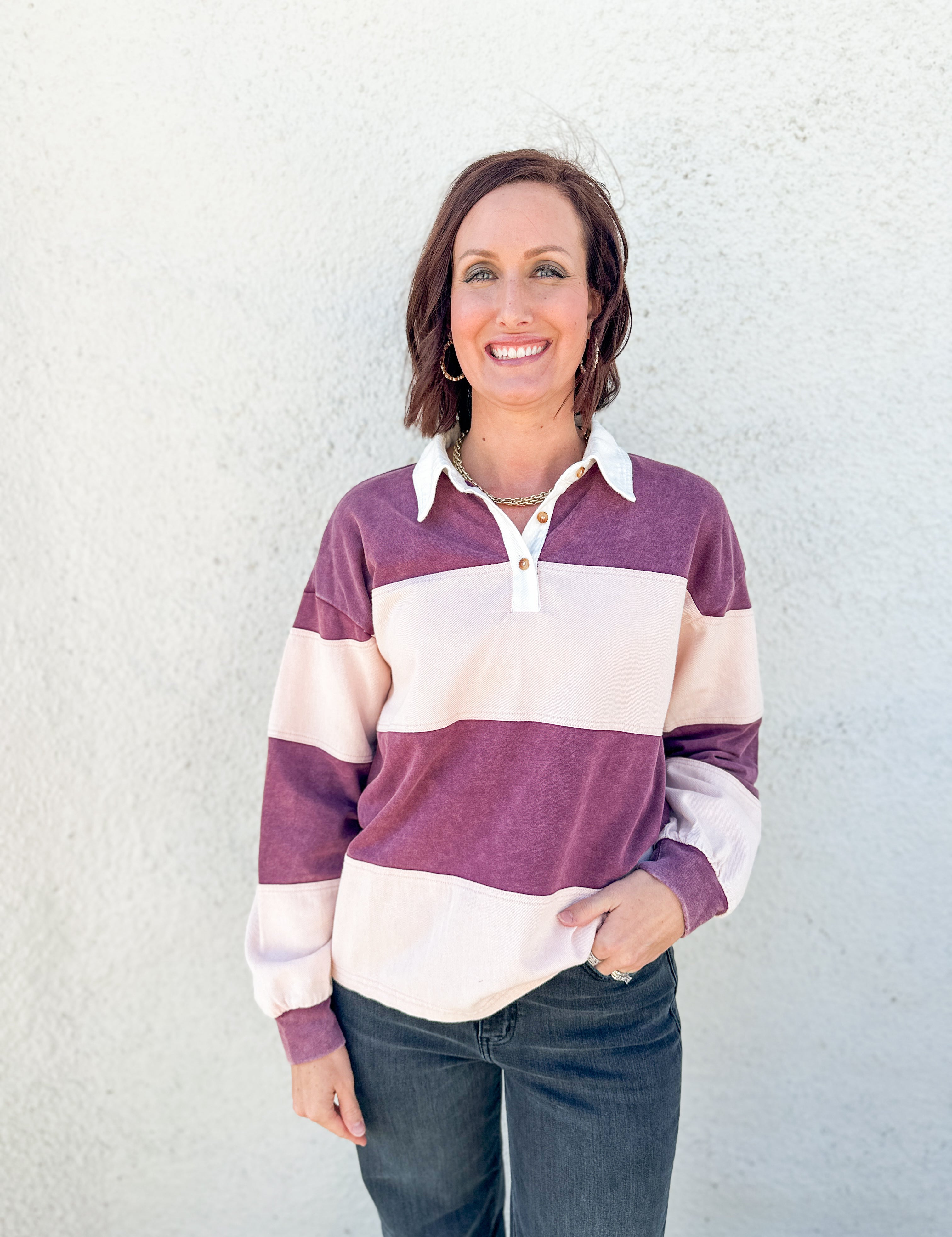 A woman smiling and wearing a long-sleeve rugby-style pullover with wide horizontal stripes in plum purple and cream, paired with dark jeans, standing against a light textured wall.