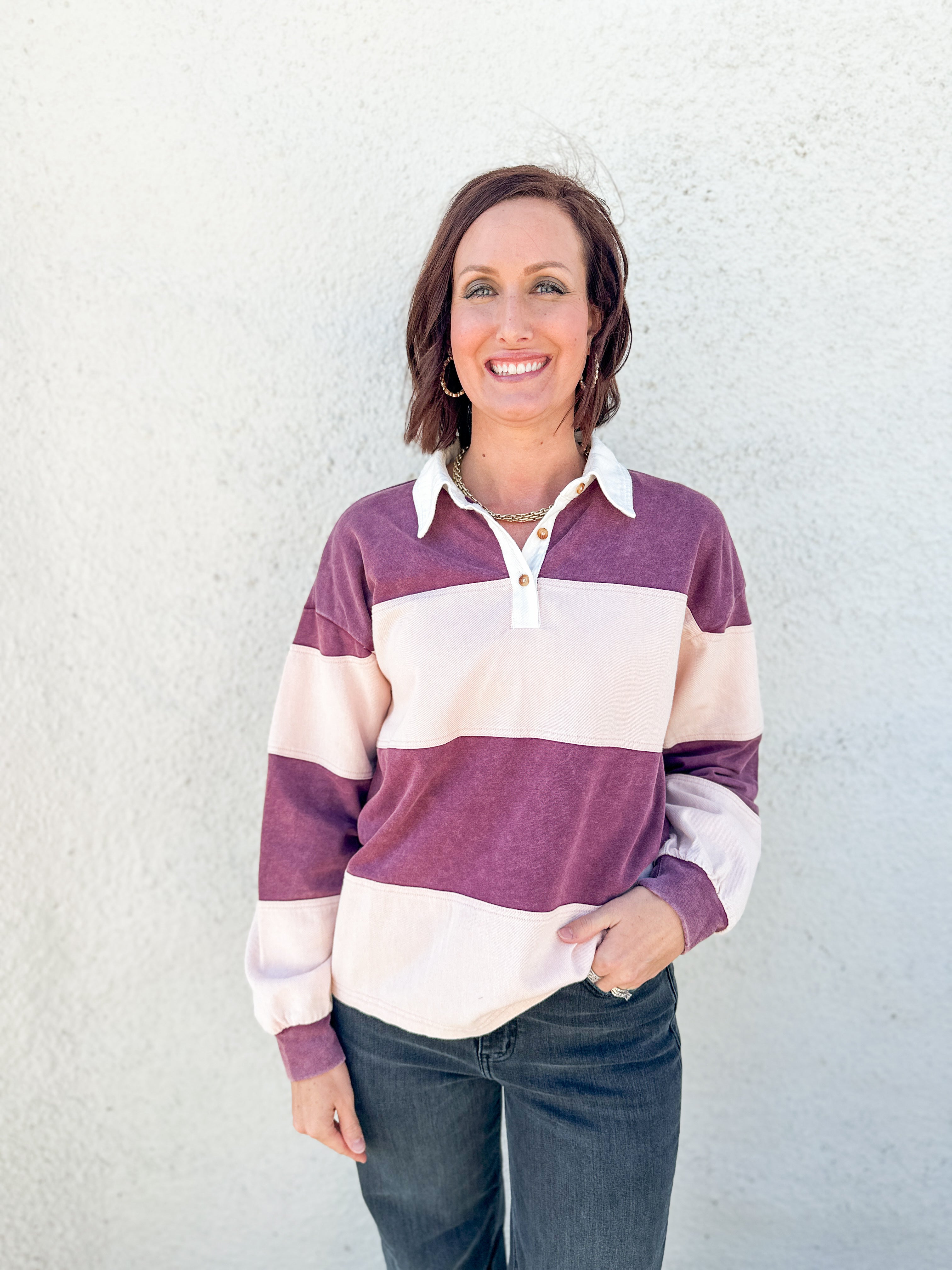 A woman smiling and wearing a long-sleeve rugby-style pullover with wide horizontal stripes in plum purple and cream, paired with dark jeans, standing against a light textured wall.
