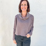 A woman with short brown hair smiling, wearing a dusty mauve cowl-neck long-sleeve top and dark gray jeans, standing against a white textured stucco wall.