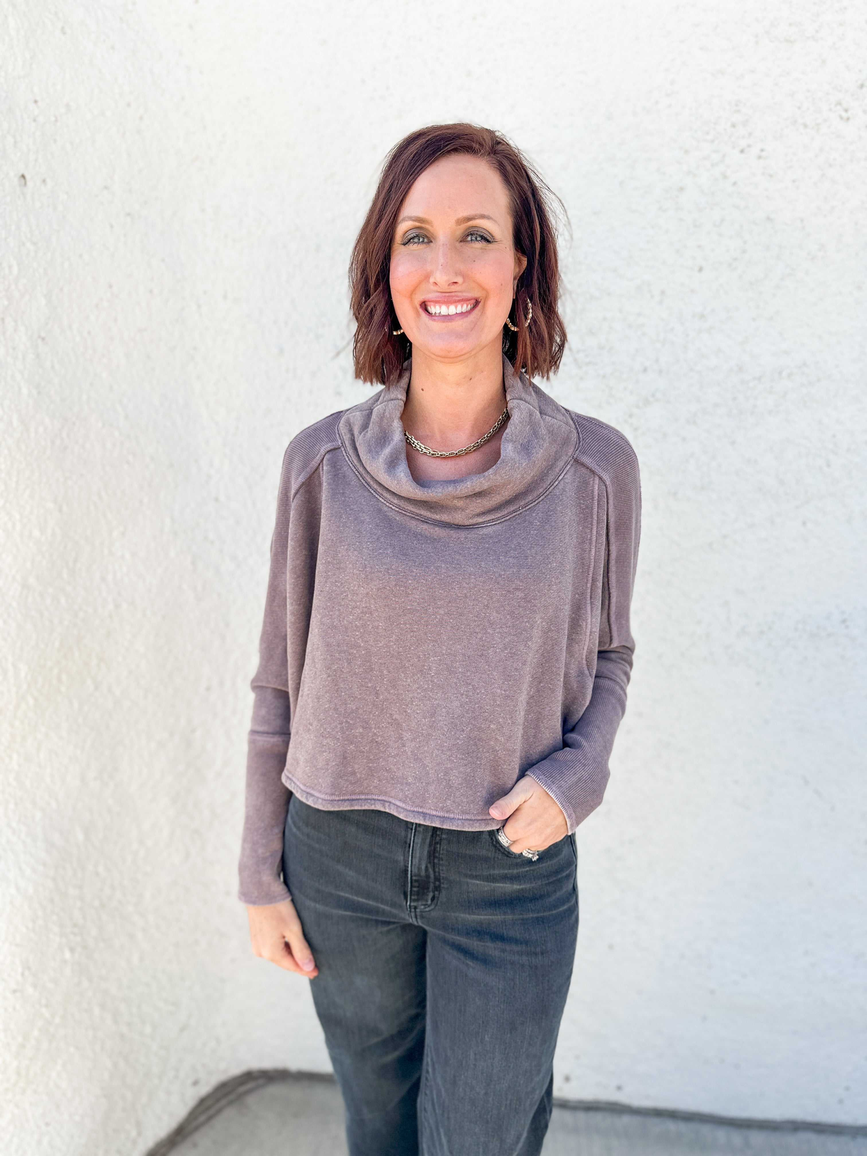 A woman with short brown hair smiling, wearing a dusty mauve cowl-neck long-sleeve top and dark gray jeans, standing against a white textured stucco wall.