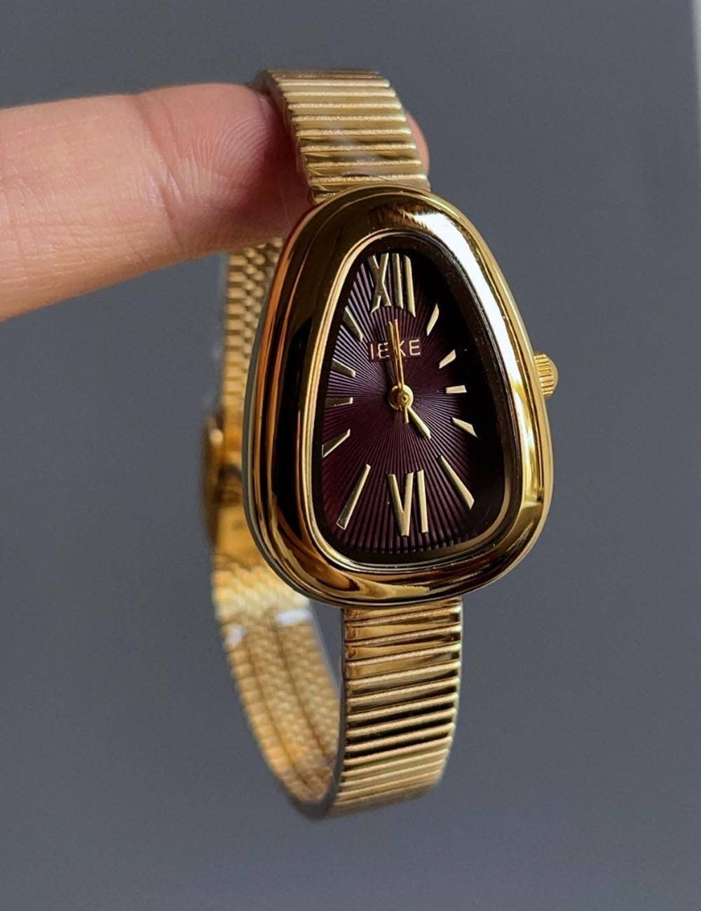 Gold watch with geometric face and burgundy face background, gold dial and roman numerals, sleek gold wristband,held by a finger against a gray background