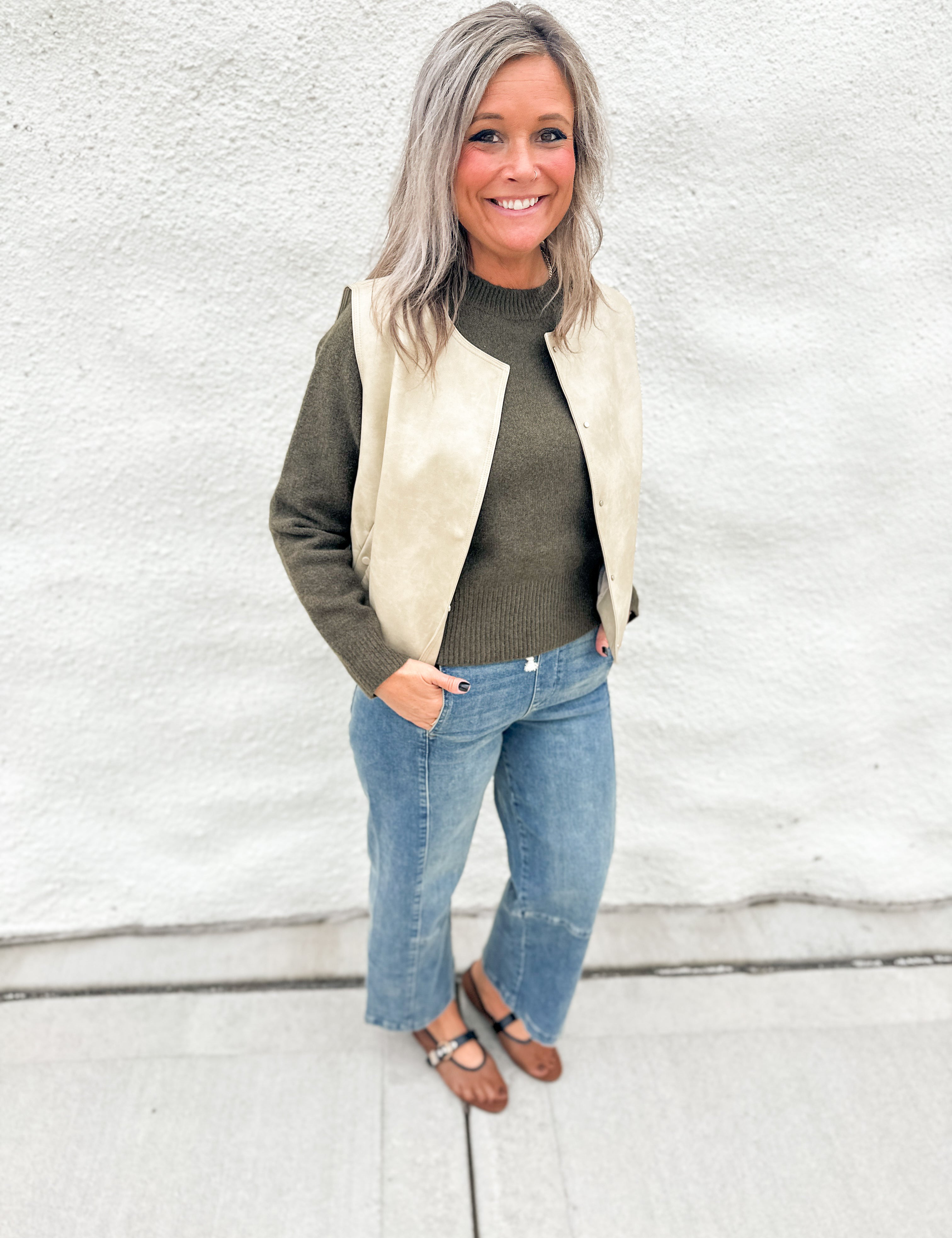 A woman with gray hair stands against a white textured wall wearing a cream faux-leather vest over an olive green sweater, blue jeans, and brown mesh Mary Jane Flats.